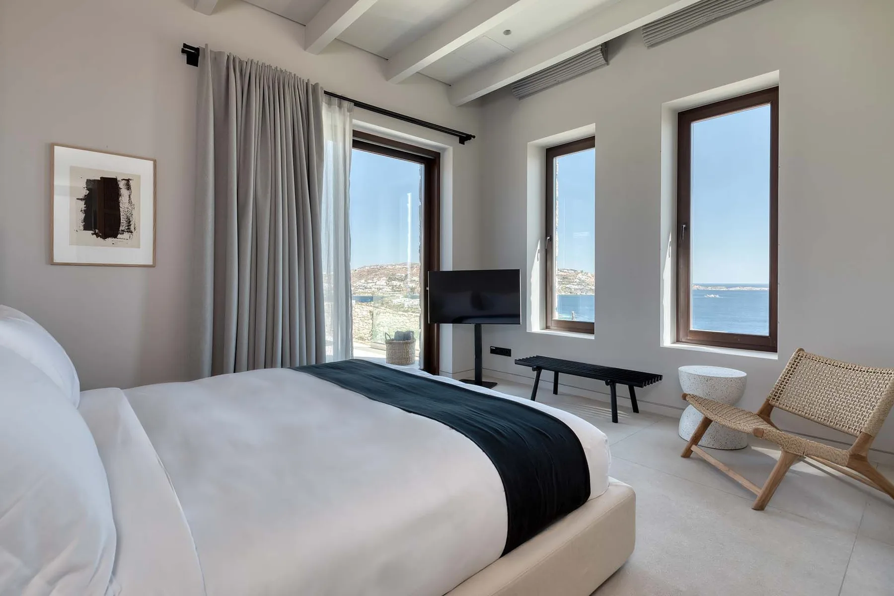A bright bedroom with a bed, television, and windows overlooking the sea.