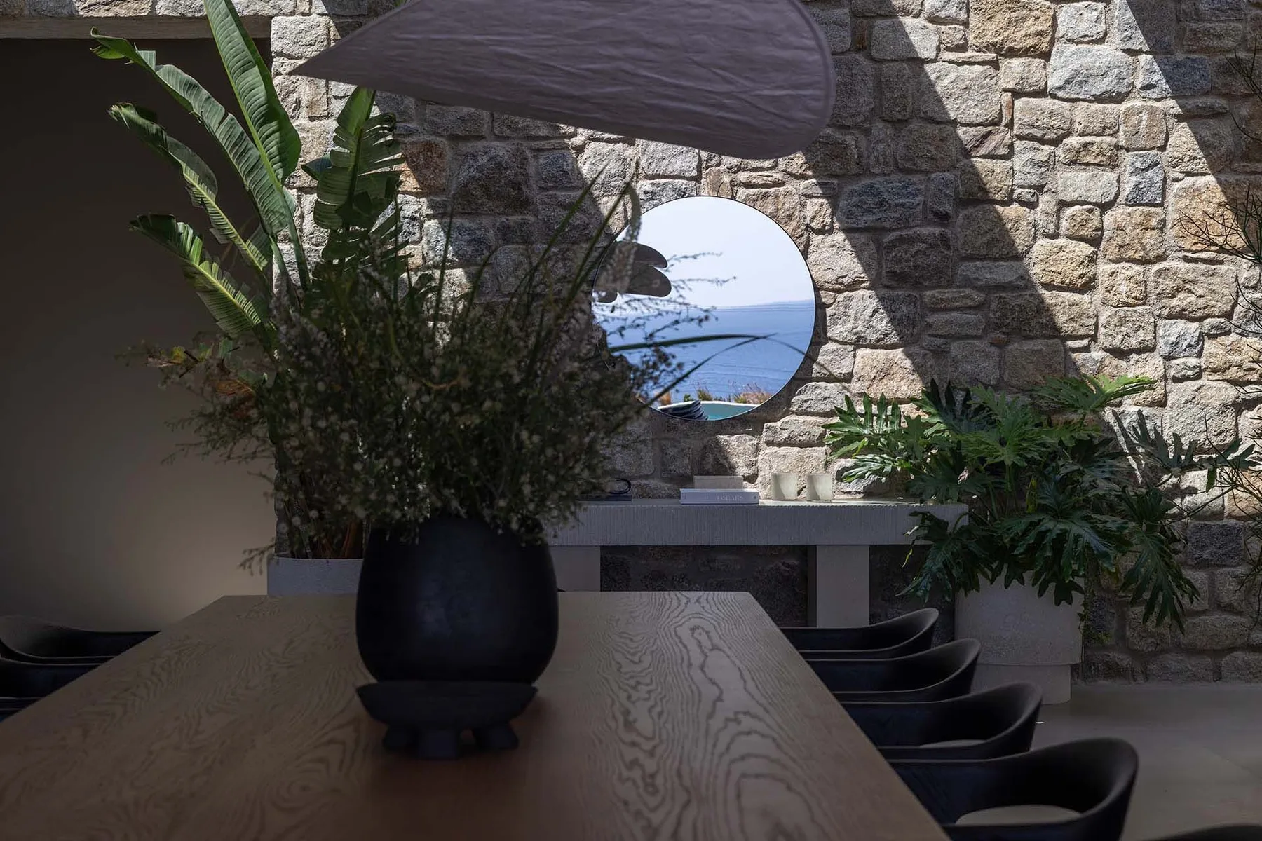 A dining area with a wooden table, dark seating, and a round mirror on the stone wall.