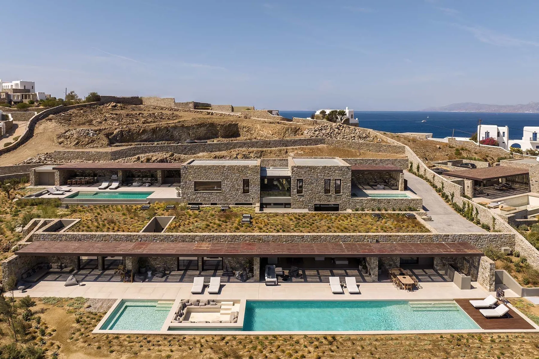 A modern resort complex with low stone buildings, landscaped terraces, and swimming pools overlooking the sea.