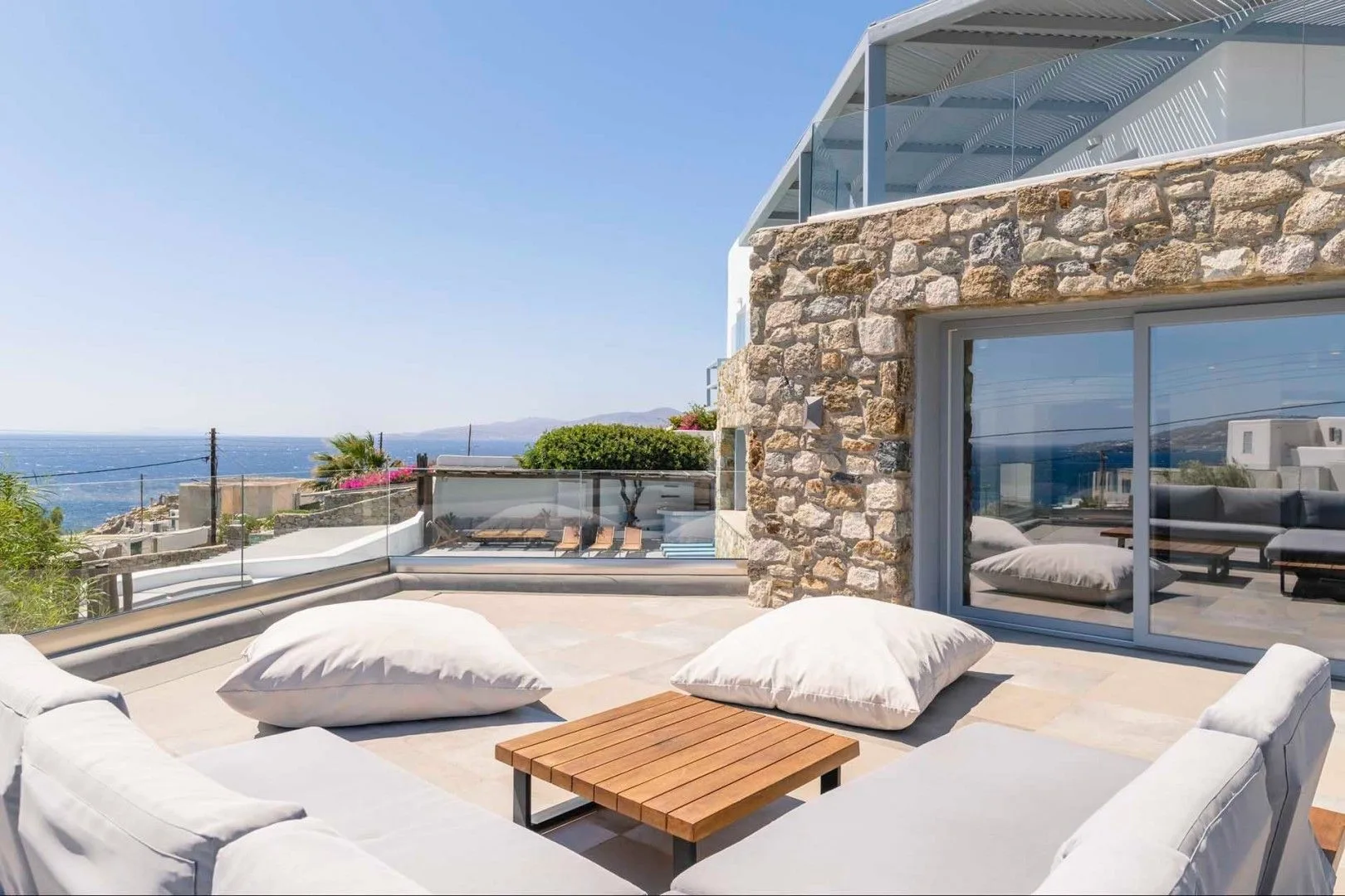 A rooftop terrace with cushioned seating and a wooden table overlooks the sea.