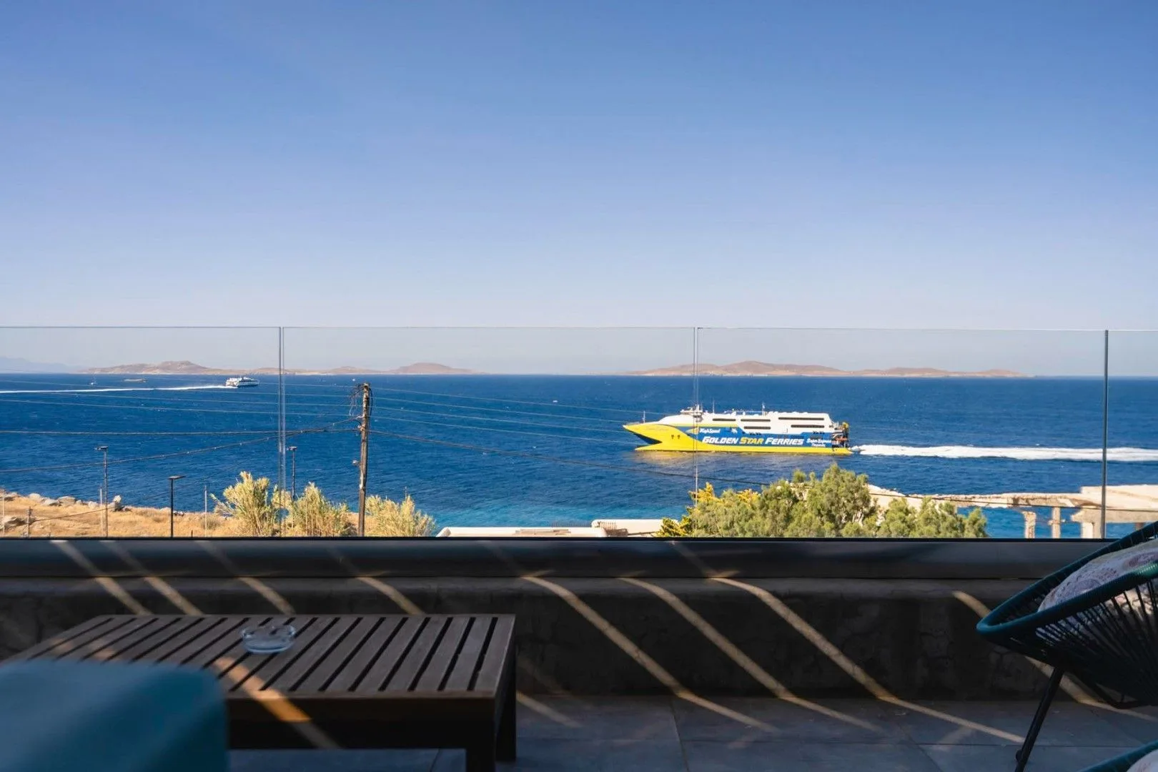 A waterfront terrace overlooks the blue sea, with a table and seating in the foreground.