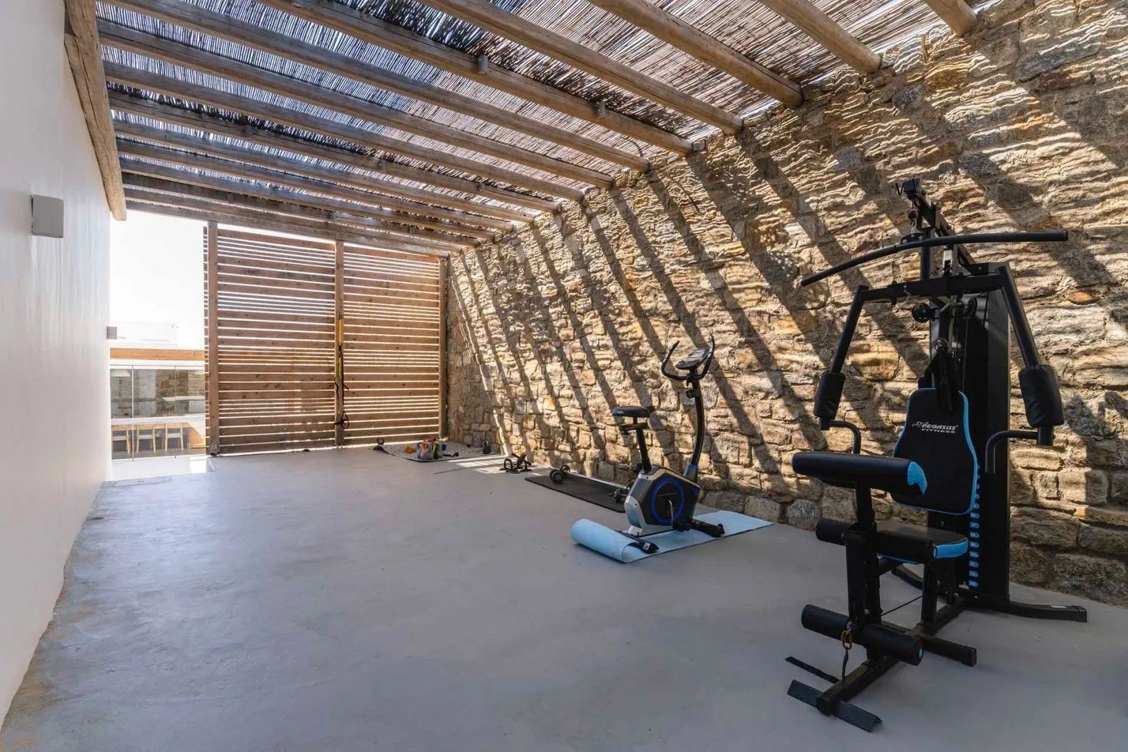 A covered exercise area has workout equipment beside a stone wall.