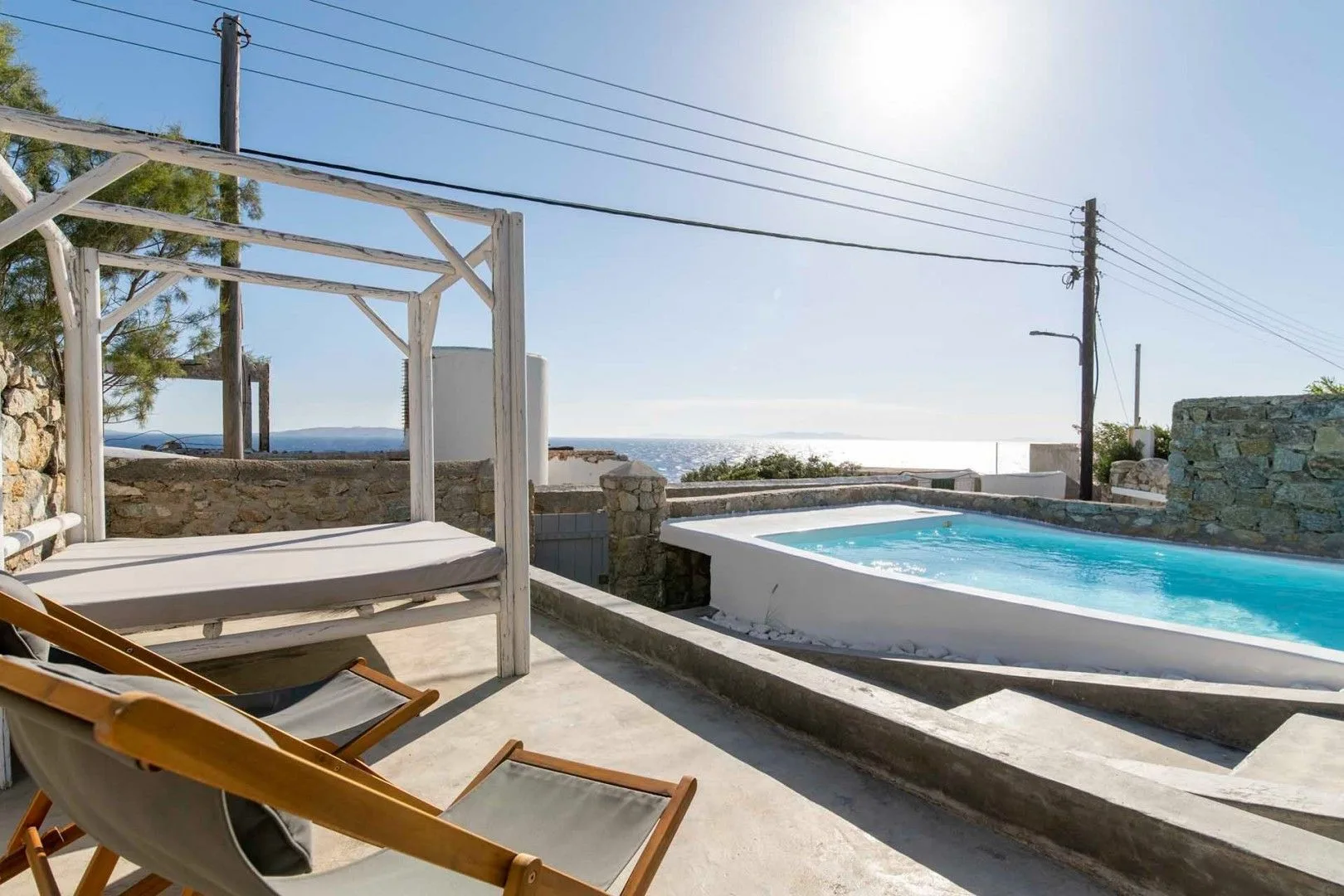 A sunlit outdoor pool beside a wooden pergola, with the sea visible beyond.