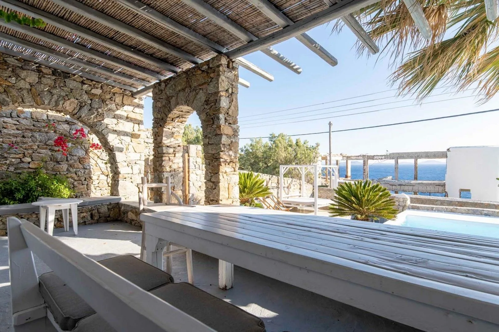 A shaded outdoor dining area with a long table and a view of the sea.