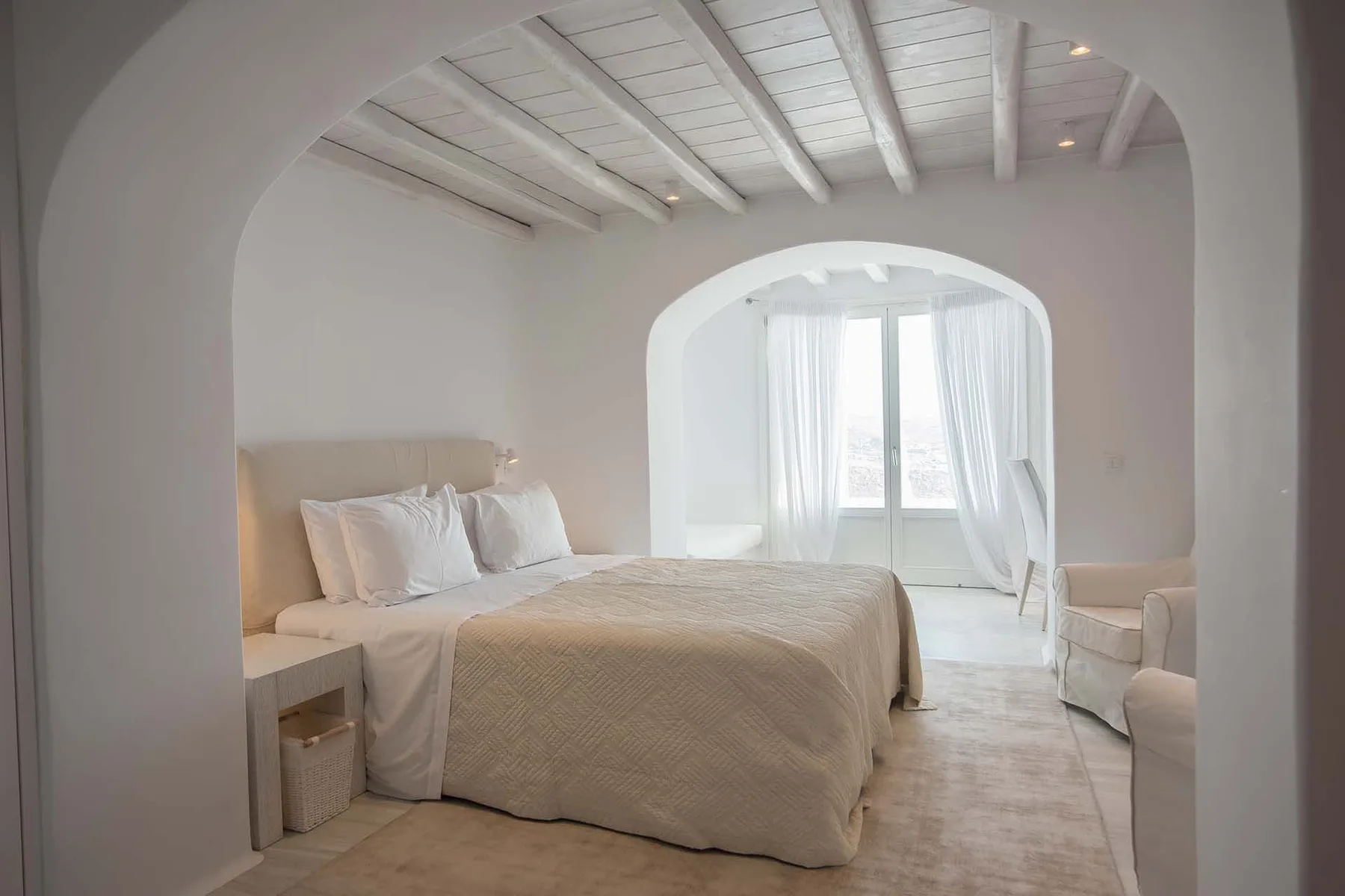 A bright bedroom with a bed beneath exposed ceiling beams and an arched opening.