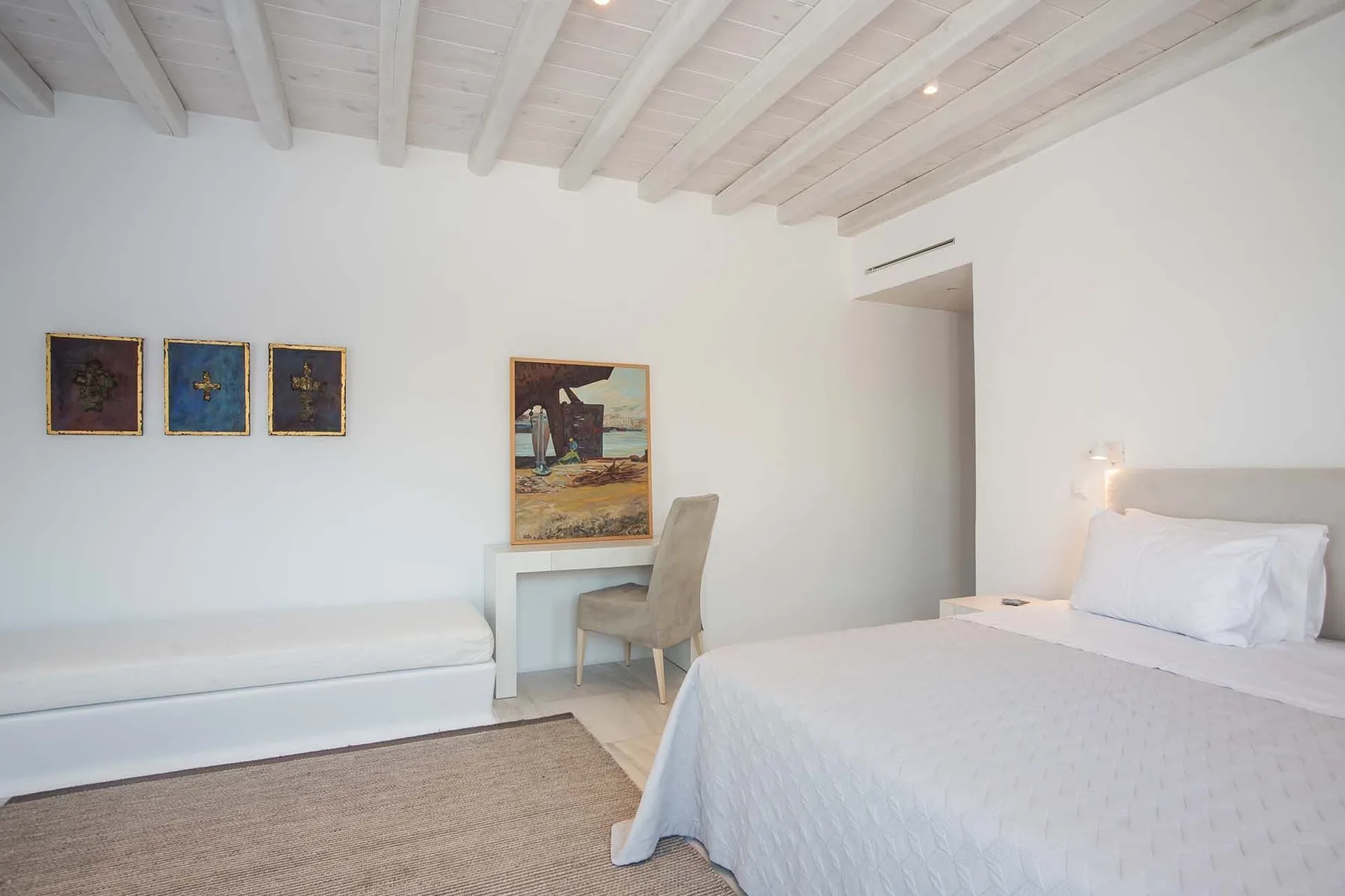A bright bedroom with a bed, a low seating area, and framed artwork.