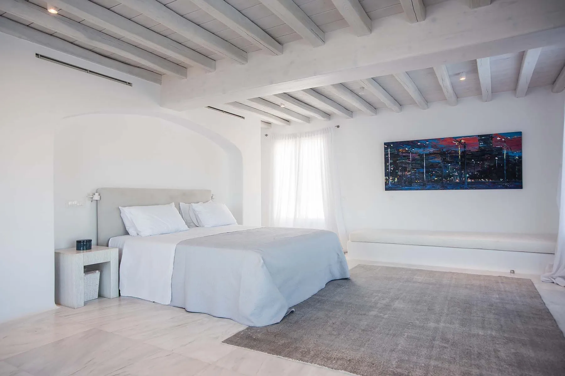 A bright bedroom with a bed beneath exposed ceiling beams and a wall painting.