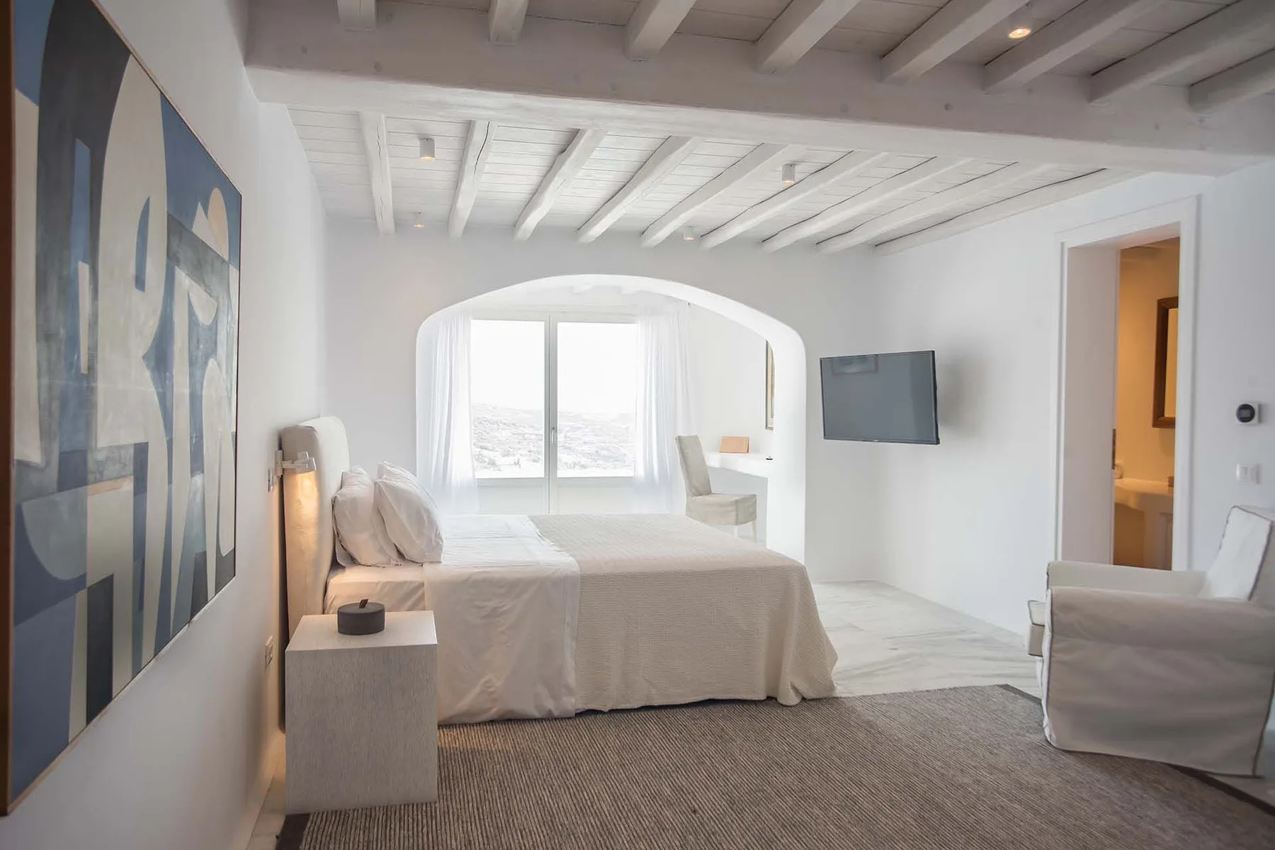 A bright bedroom with a bed beneath a white beamed ceiling and an arched window.