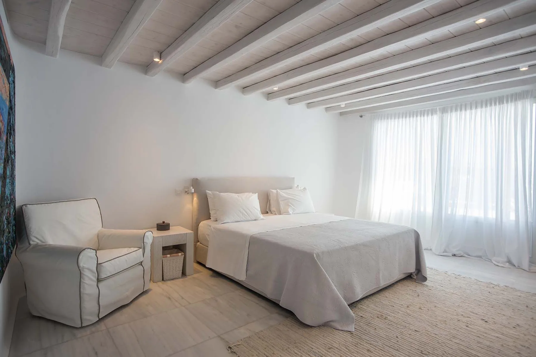 A bright bedroom with a bed, an armchair, and sheer curtains.