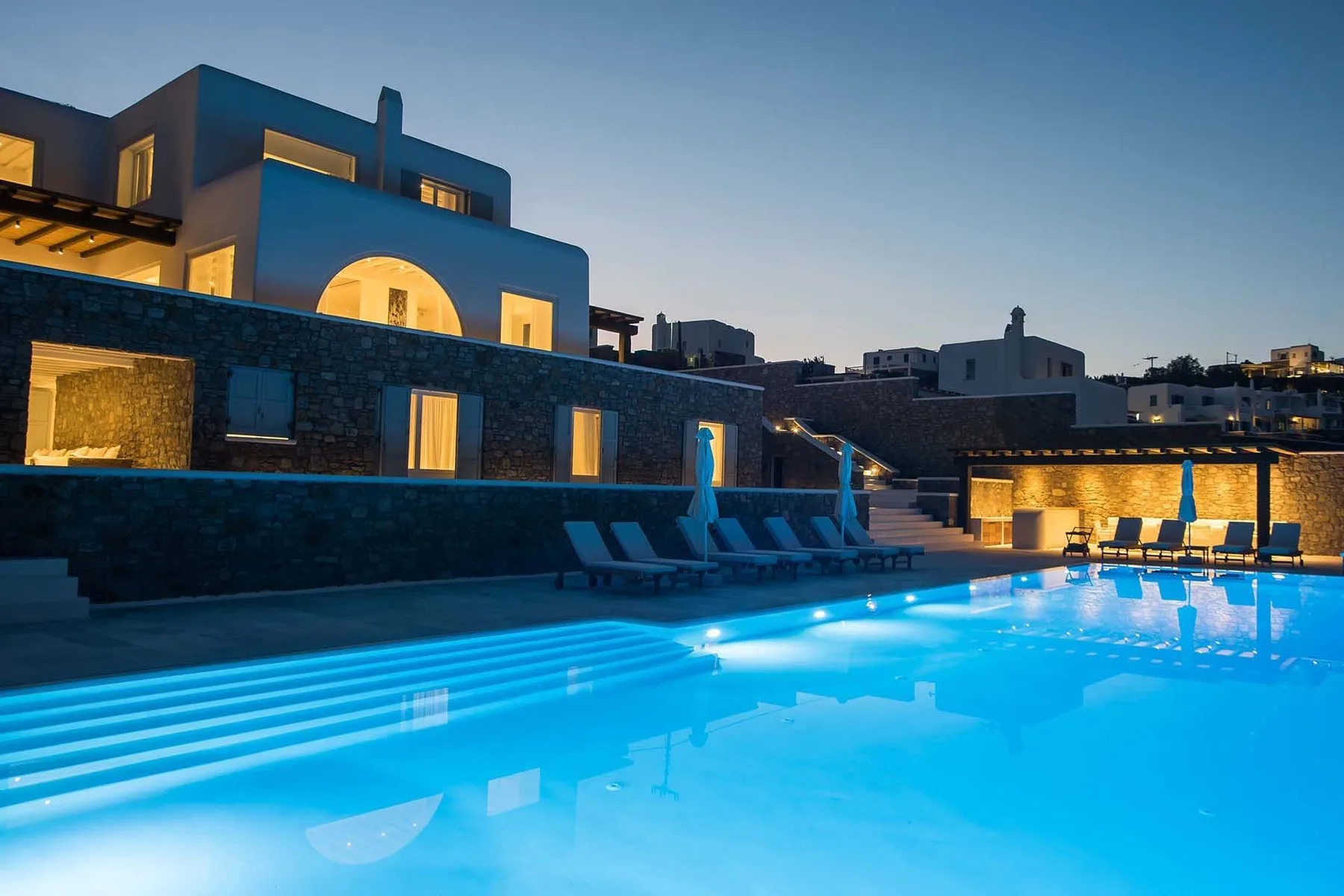 A lit swimming pool sits beside modern buildings at dusk.