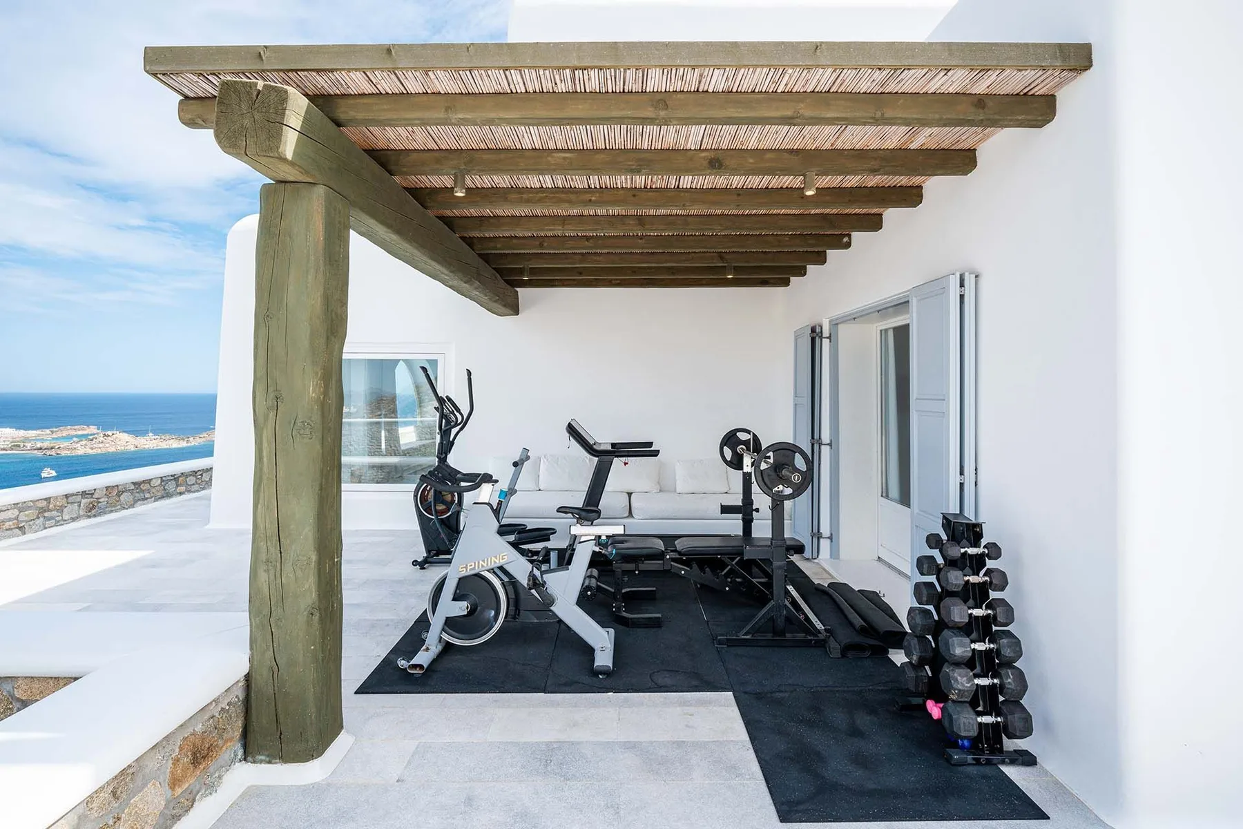 An outdoor gym with exercise equipment beneath a wooden pergola beside the sea.