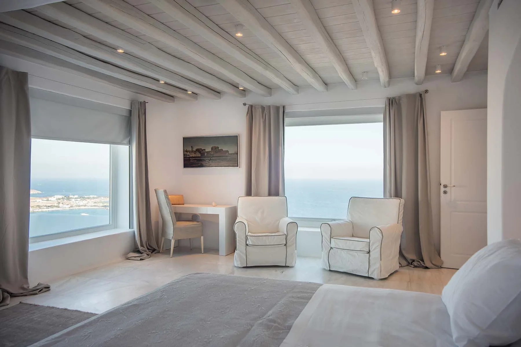A bright bedroom with two armchairs facing large windows overlooking the sea.