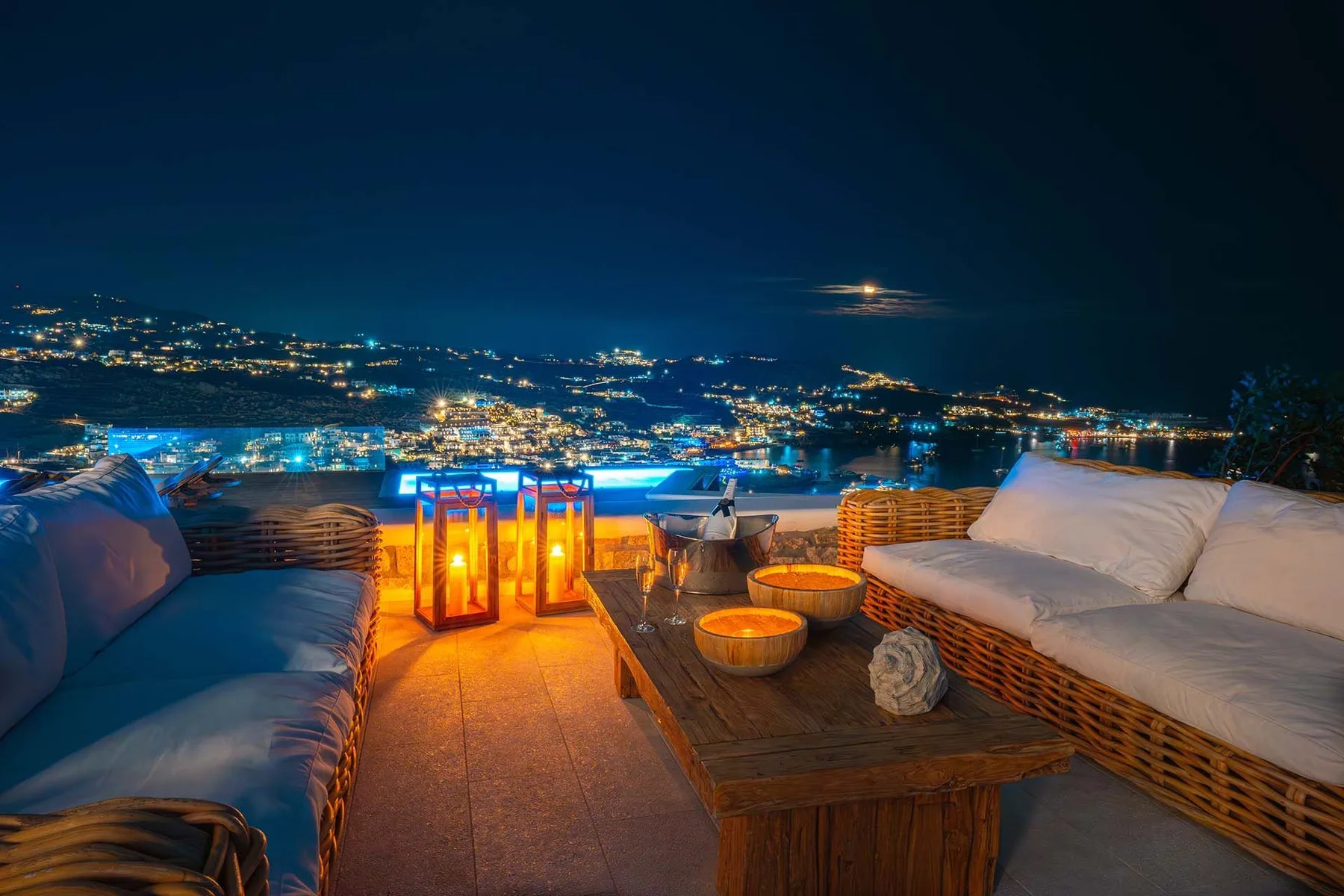 An outdoor terrace at night overlooks a city, with cushioned seating and lanterns.