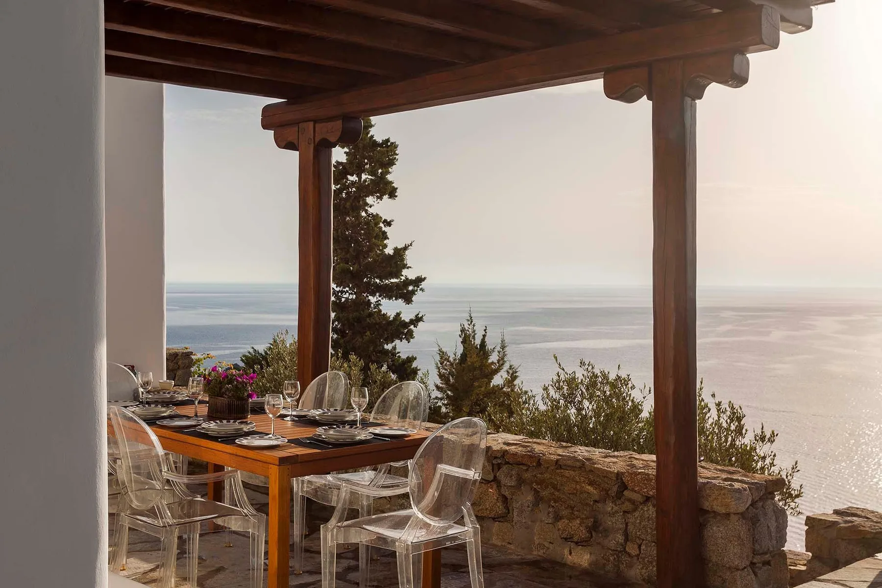 A dining table with clear seating sits on a covered terrace overlooking the sea.
