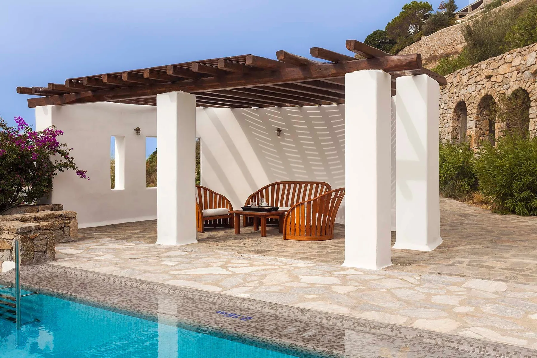 A shaded outdoor seating area beside a swimming pool, with a wooden pergola overhead.