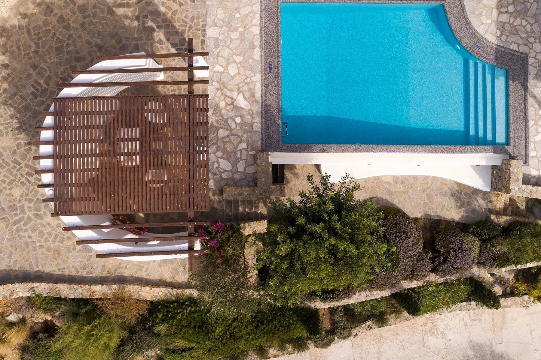 An outdoor swimming pool beside a wooden slatted pergola on a stone patio.