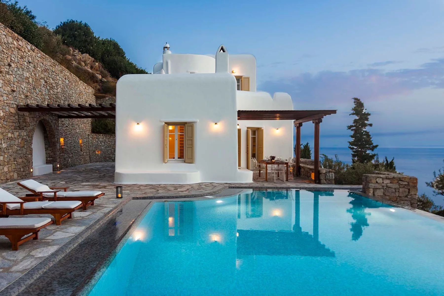 A white seaside villa overlooks a swimming pool and the ocean.