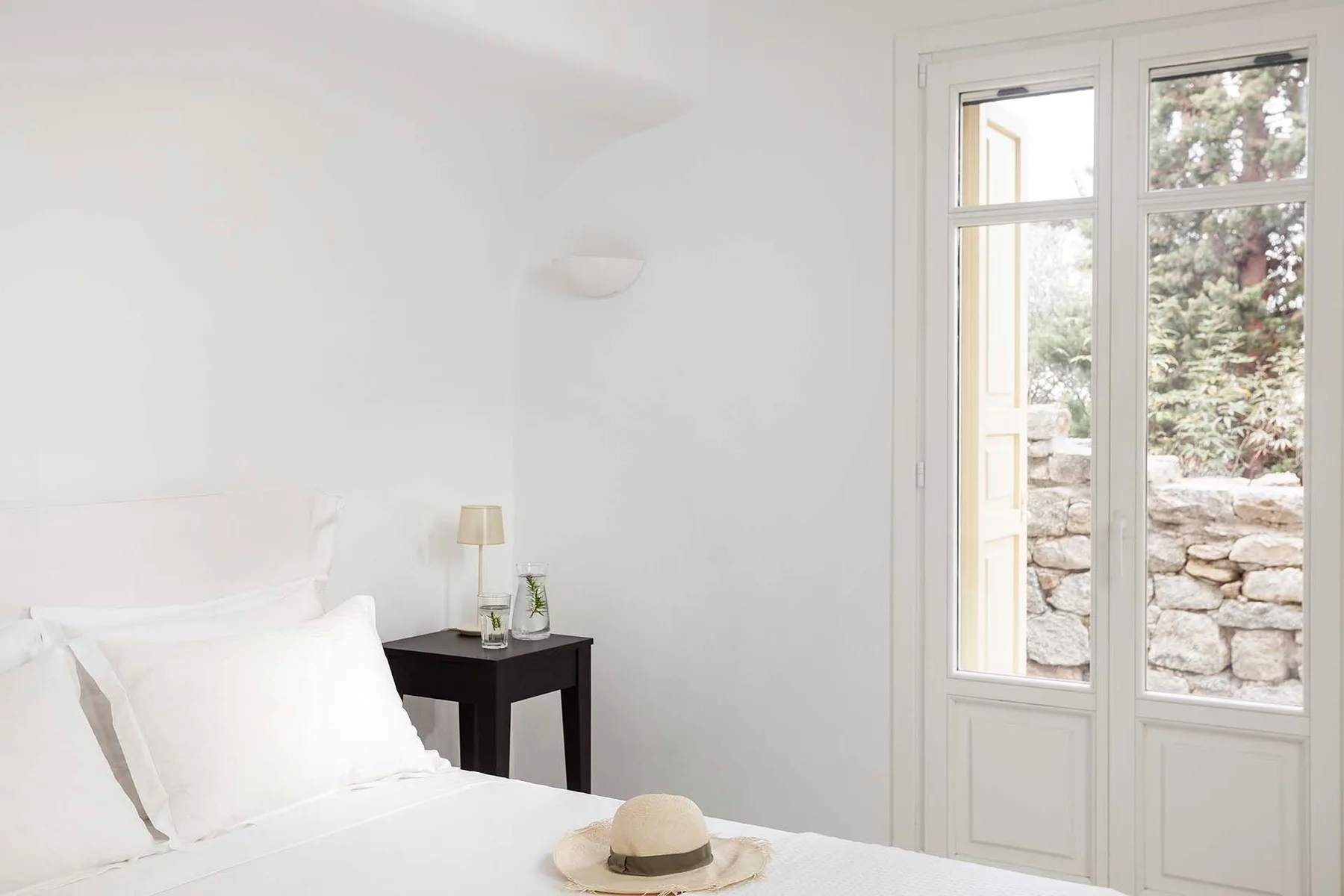 A bright bedroom with white bedding and tall glass doors overlooking a stone wall.