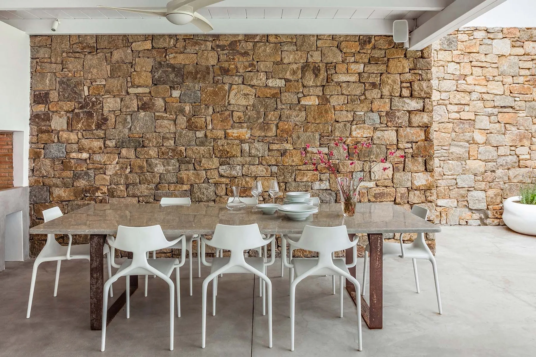A dining area with a long table and white seating in front of a stone wall.