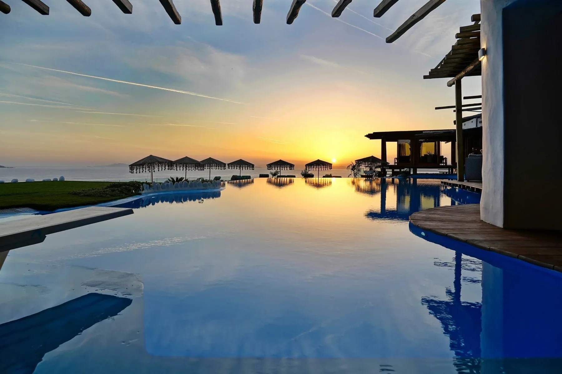 A swimming pool reflects the sunset beside the sea and shaded beach seating.