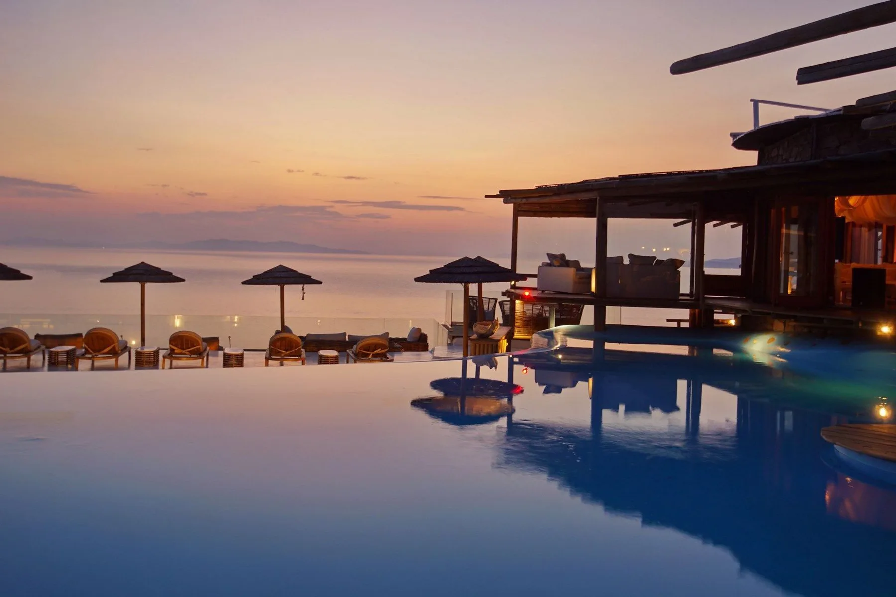An infinity pool overlooks a calm waterfront at sunset, with shaded seating nearby.
