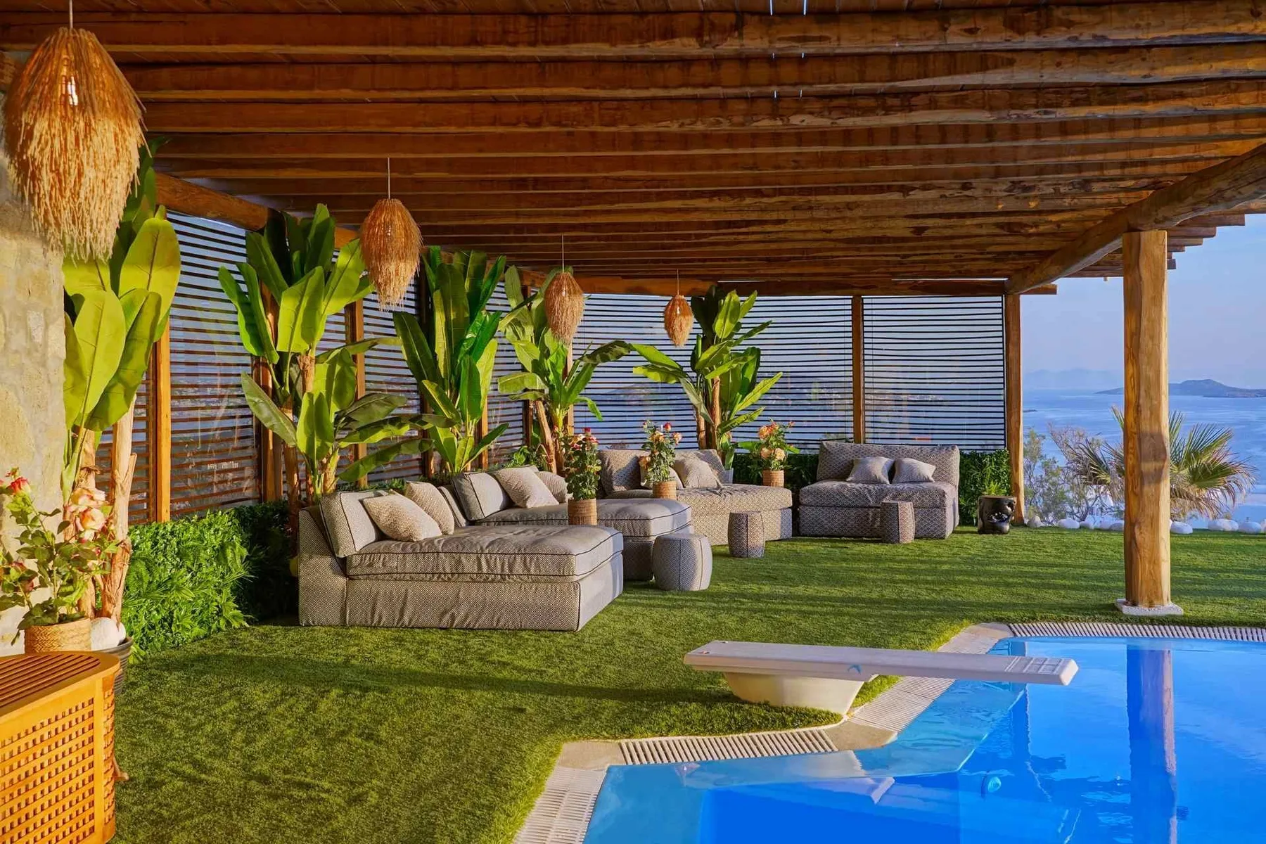 A covered outdoor seating area beside a swimming pool, with plants and cushioned furniture.