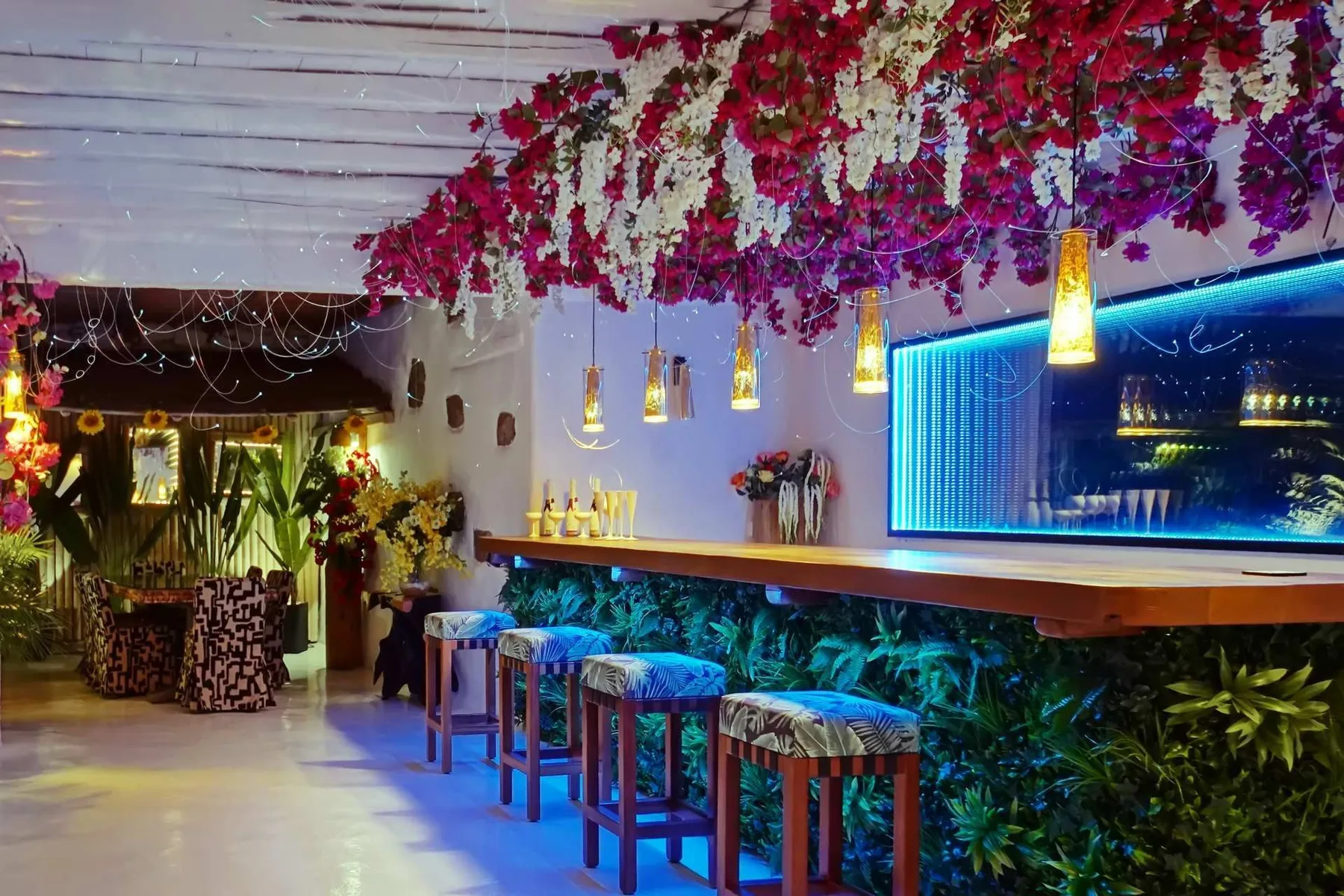 A bar counter with stools sits beneath hanging flowers and pendant lights.