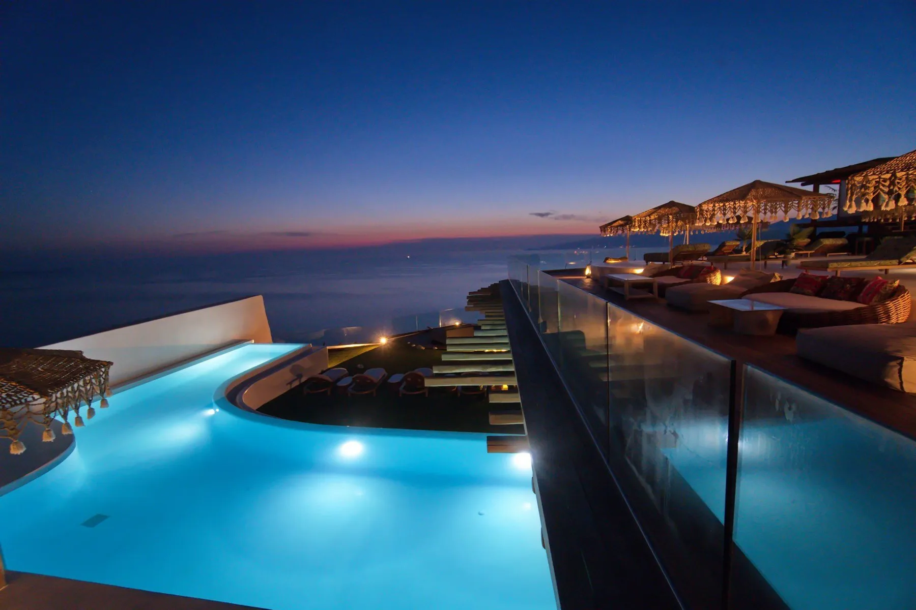 An illuminated swimming pool overlooks the sea at dusk, beside an outdoor seating area.