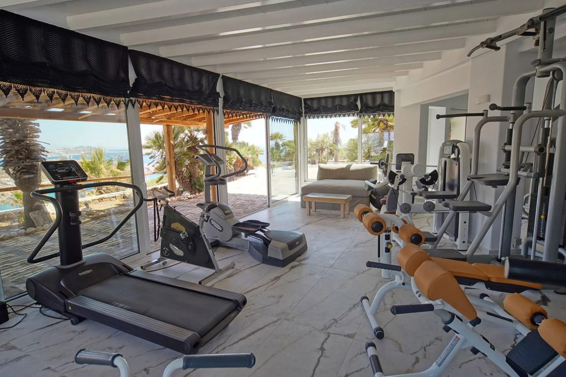 A bright home gym with exercise machines beside large windows overlooking palm trees.