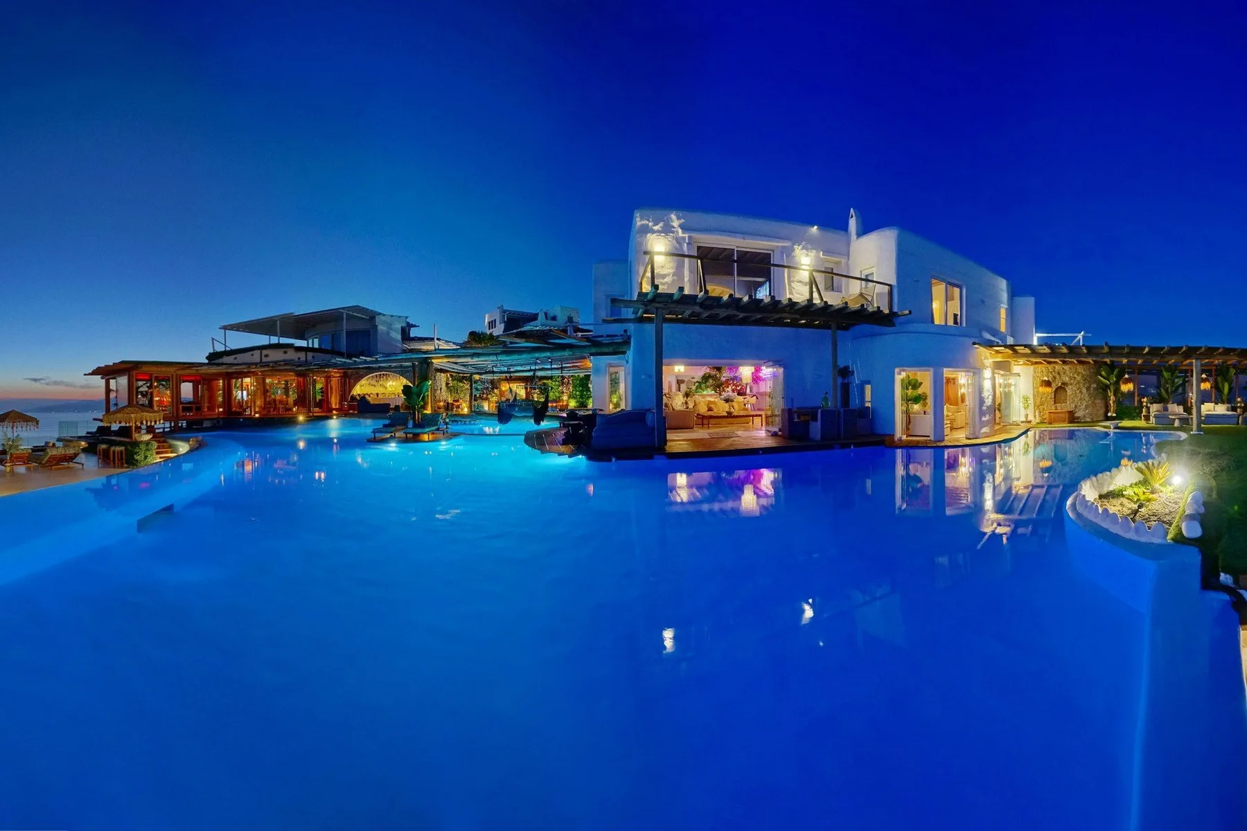 A large illuminated swimming pool reflects modern buildings at dusk.