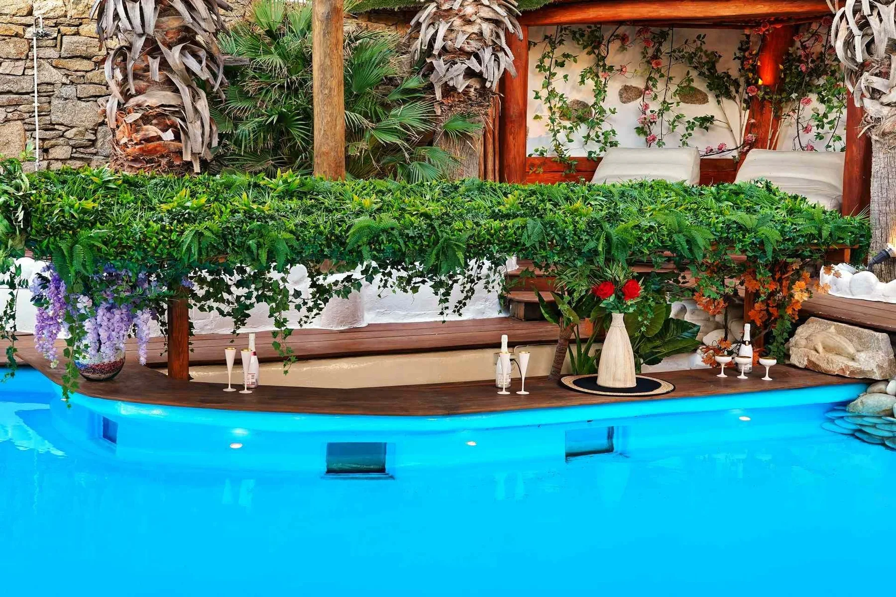 A bright blue swimming pool sits beside a garden patio with leafy plants.