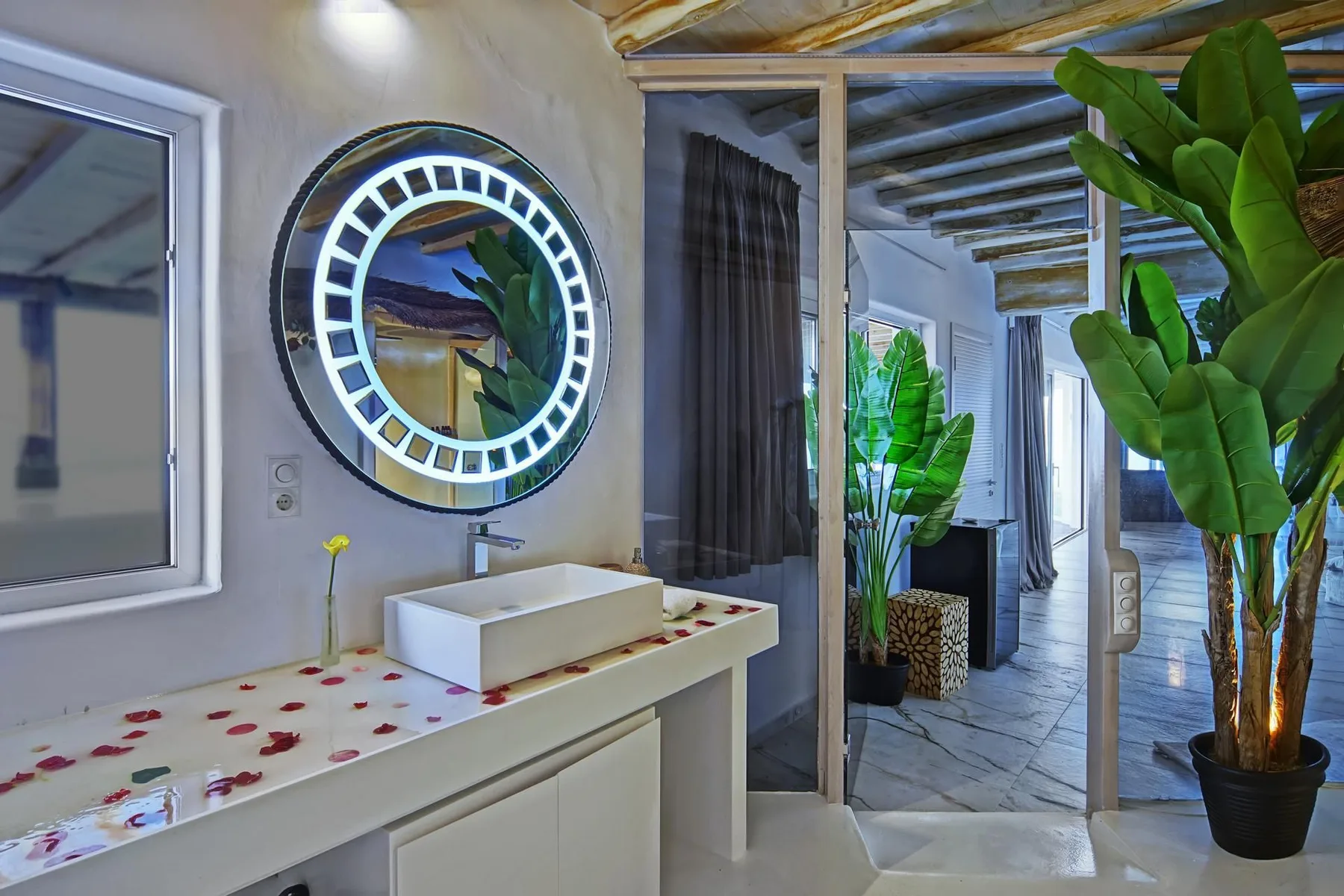 A bathroom vanity with a vessel sink and illuminated round mirror opens onto a plant-filled interior.