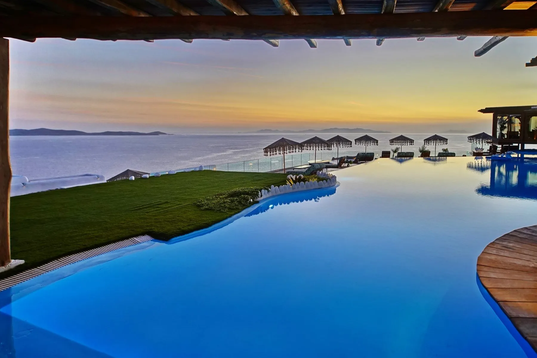An infinity pool overlooks the sea at sunset, with a grassy area beside it.