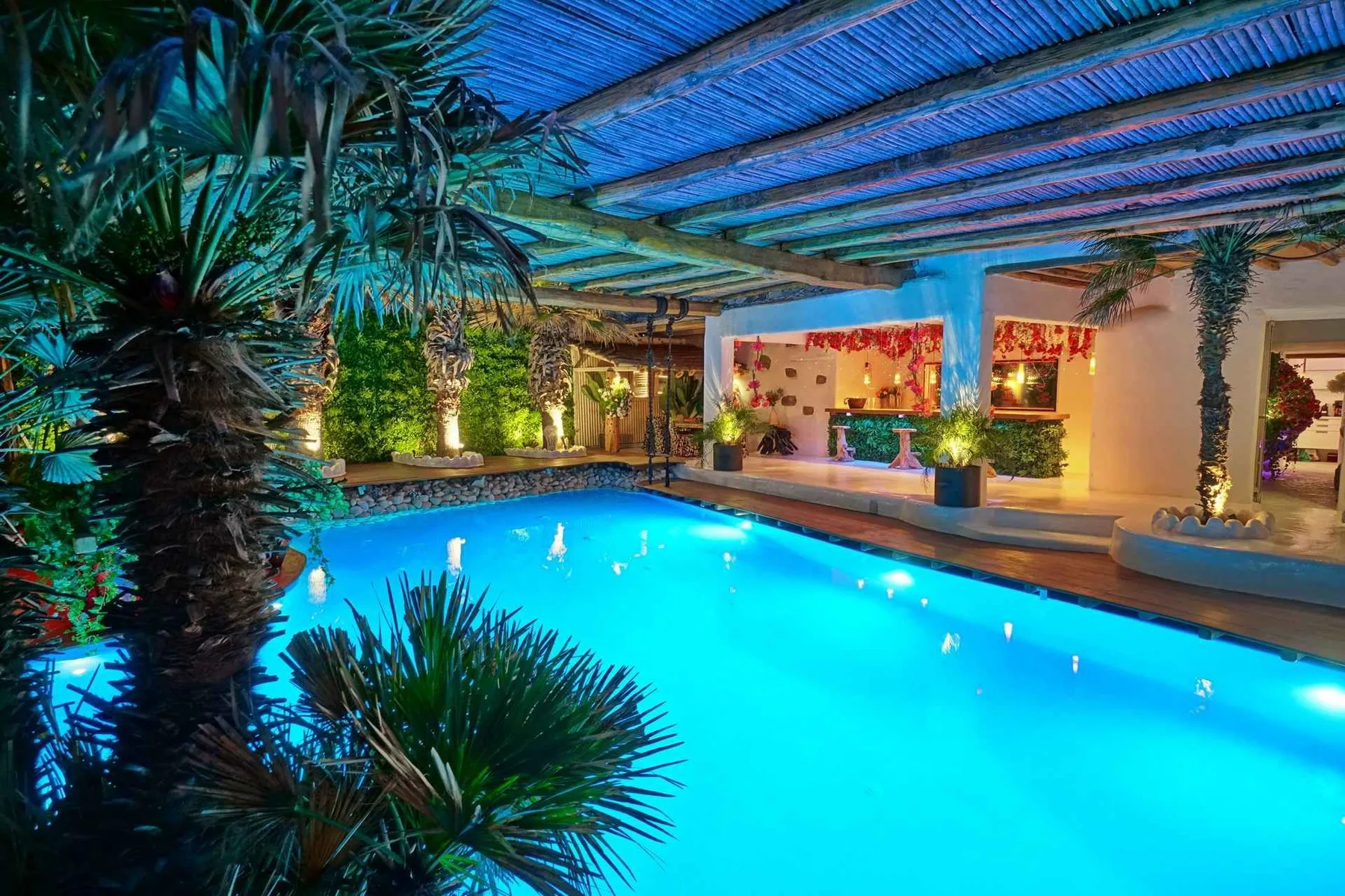 A brightly lit swimming pool sits beneath a covered patio surrounded by tropical plants.