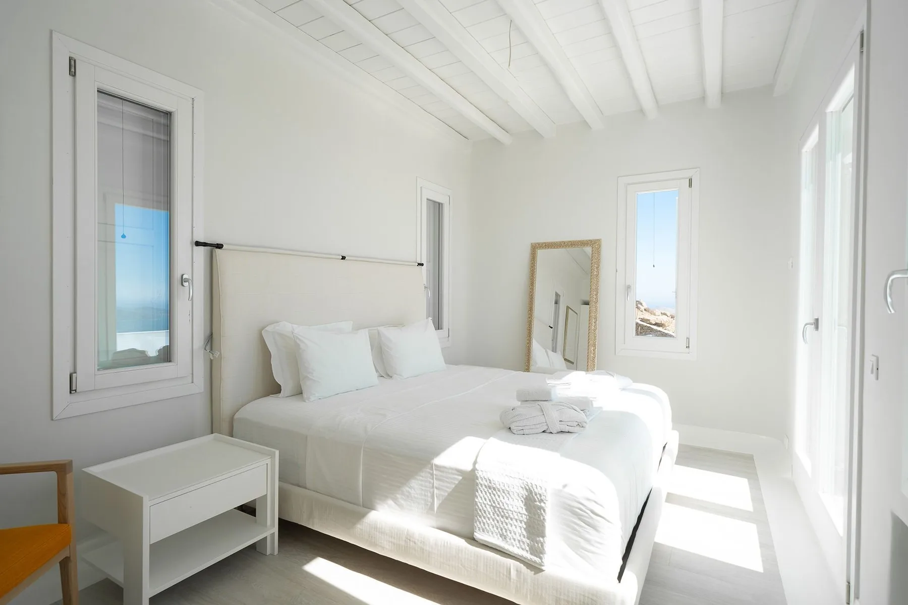 A bright bedroom with a white bed beneath a beamed ceiling.