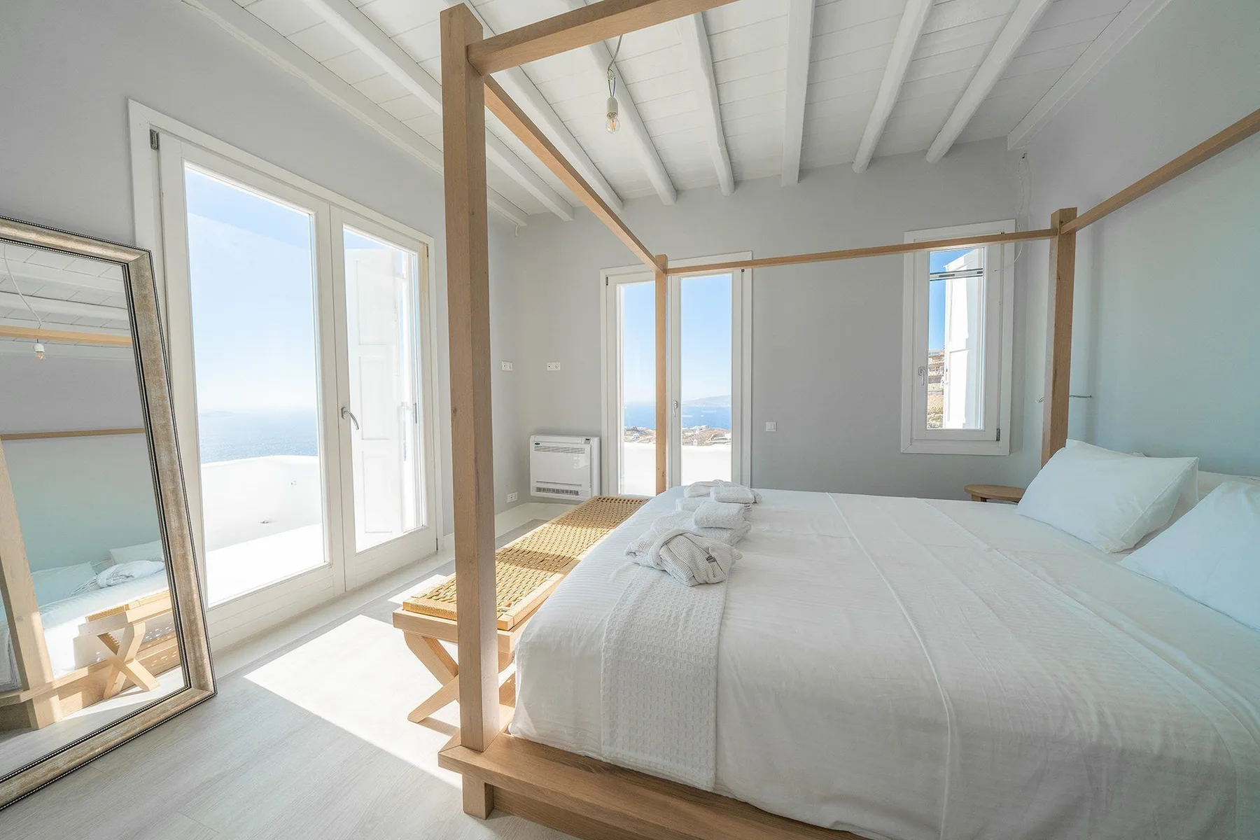 A bright bedroom with a large bed, wooden frame, and windows overlooking the sea.