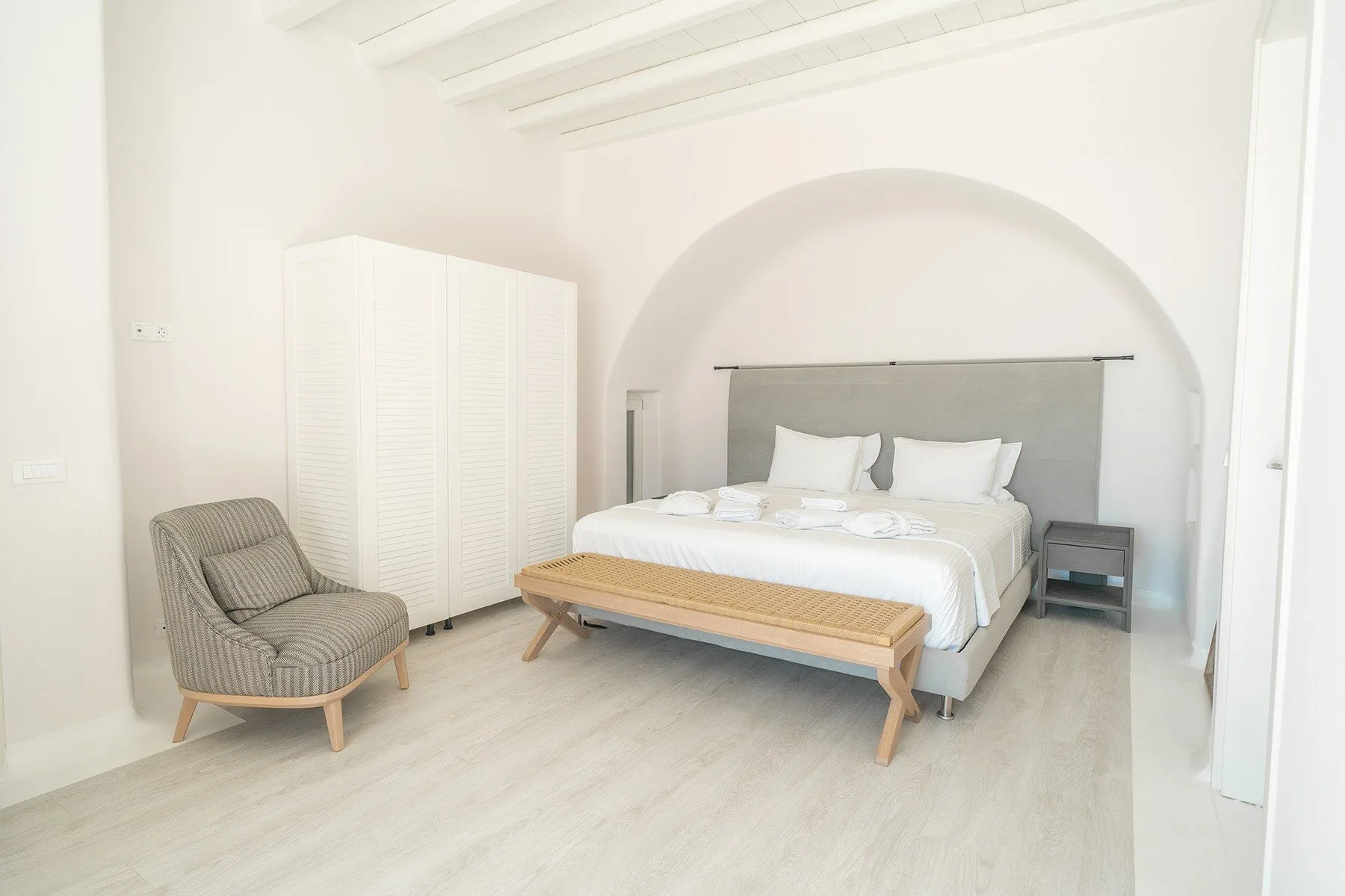 A bright bedroom with a bed, an upholstered chair, and a white wardrobe.