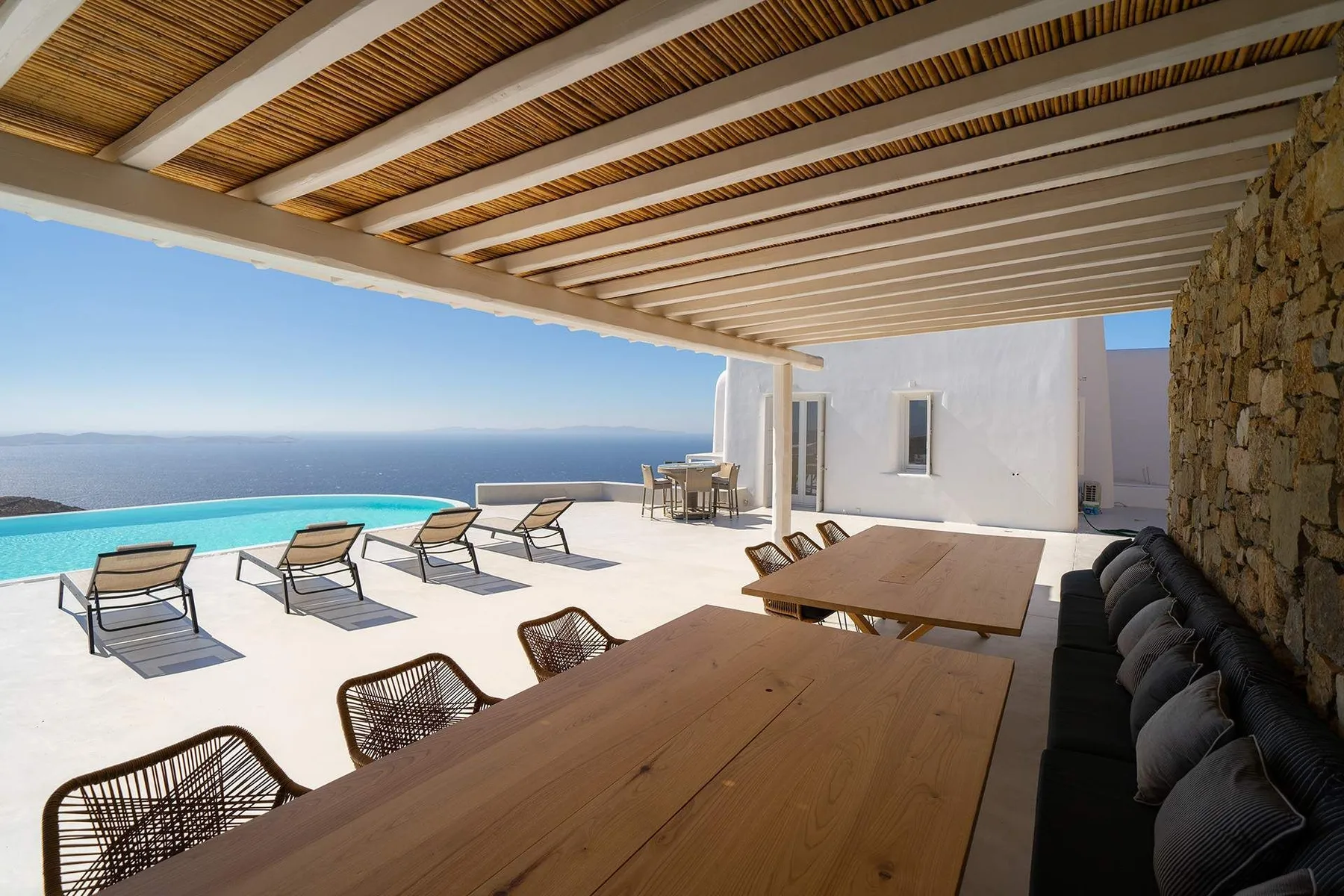 An outdoor terrace with dining tables and lounge seating overlooks a pool and the sea.