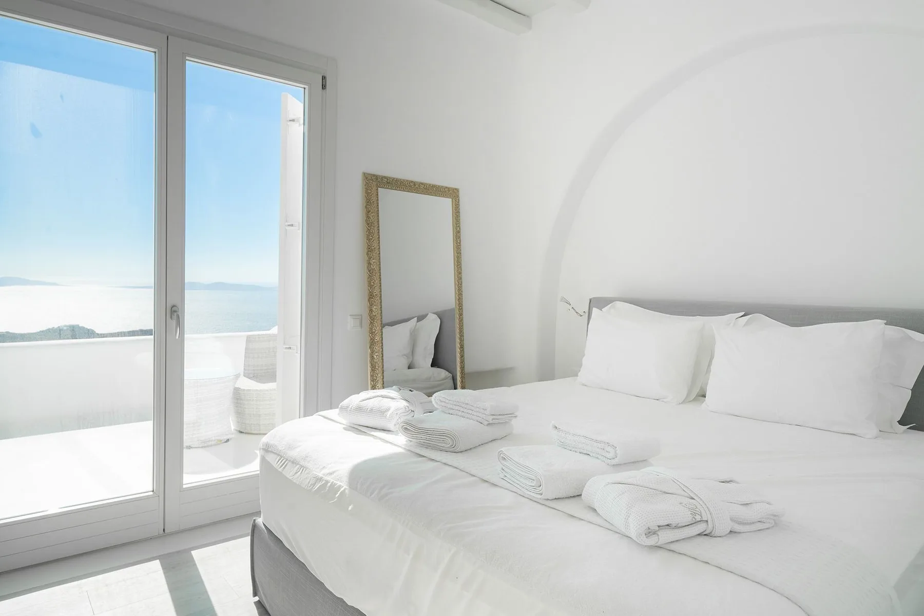 A bright bedroom with white bedding beside large glass doors overlooking the water.