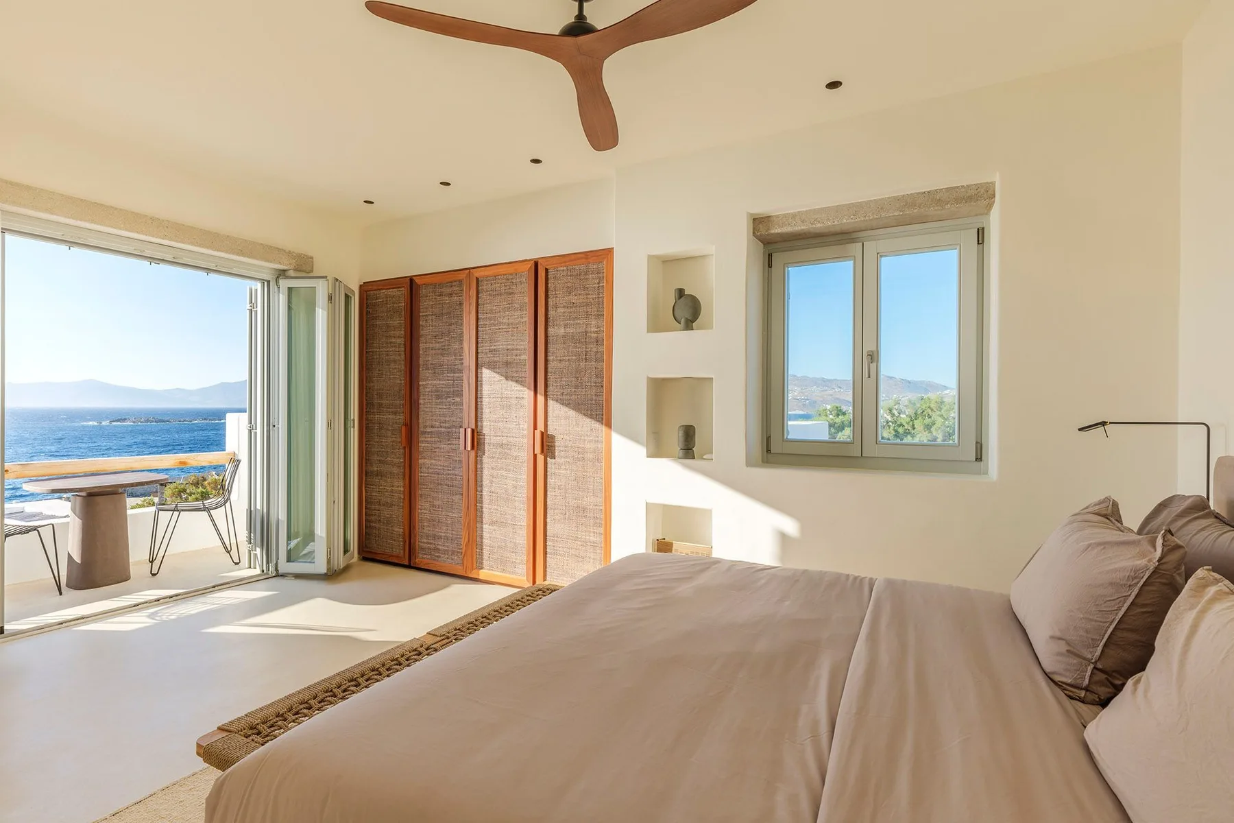 A bright bedroom with a bed, open balcony doors, and a view of the sea.