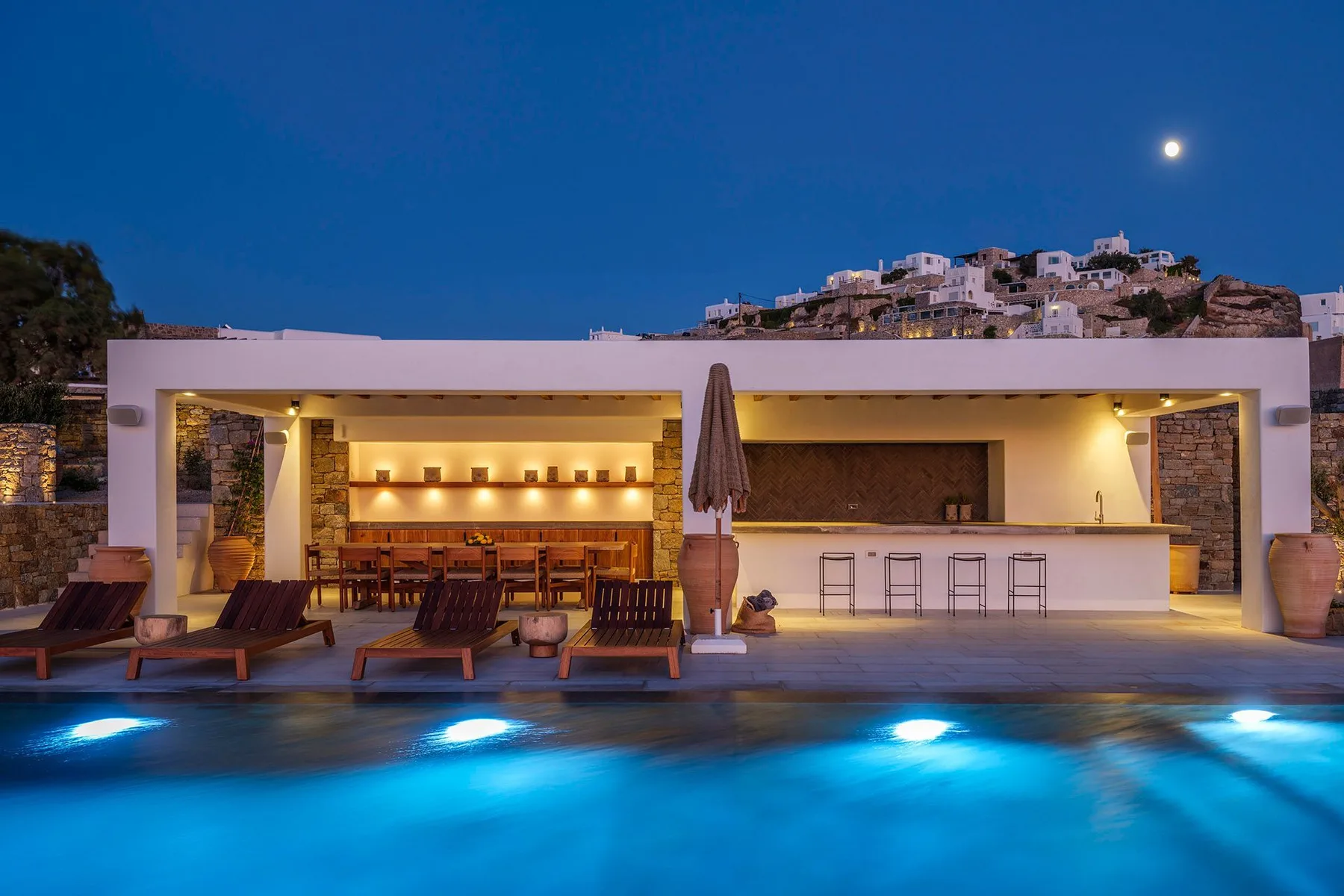 An illuminated poolside bar and seating area overlook a glowing swimming pool at dusk.