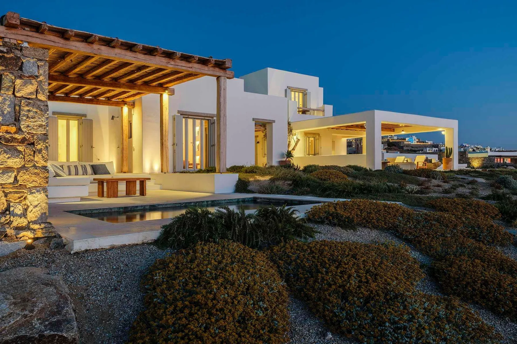 A modern white house with a warmly lit patio and landscaped garden at dusk.