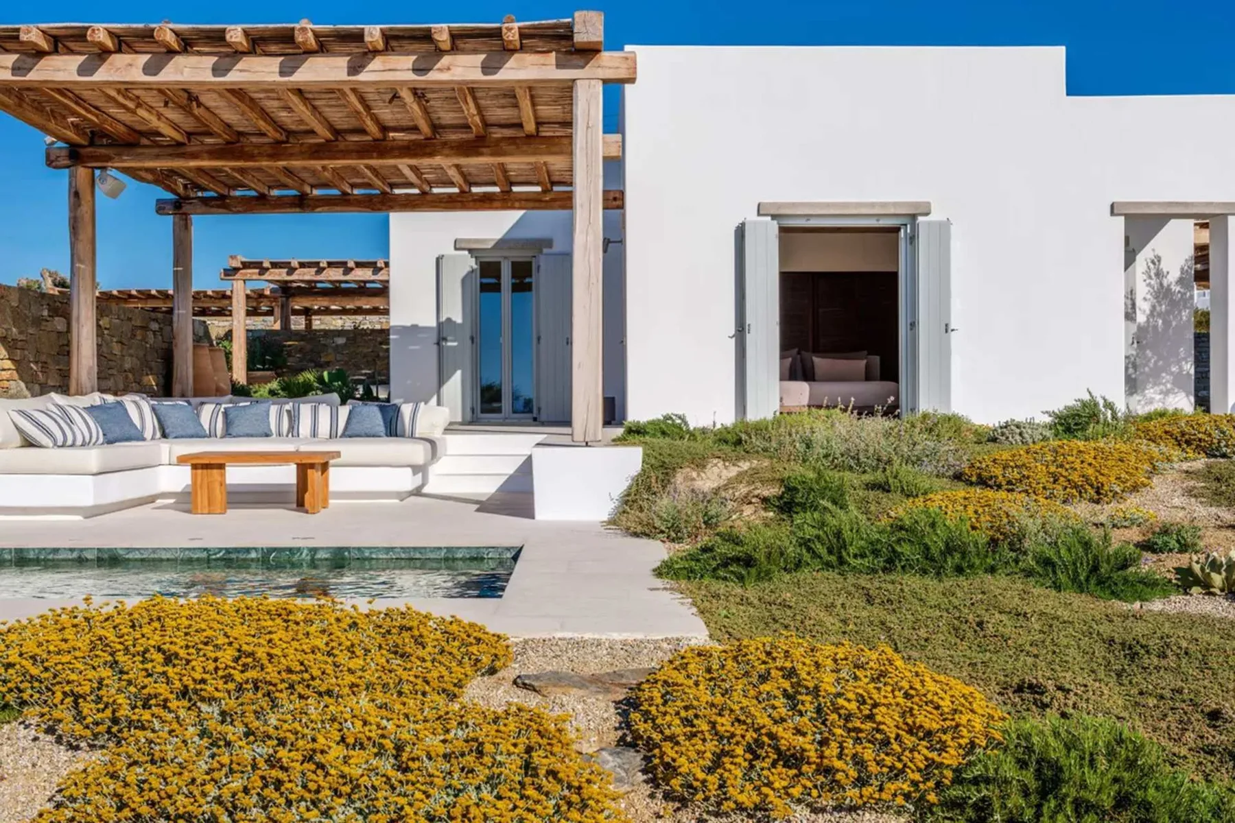 A white modern house opens onto a patio with a wooden pergola and swimming pool.