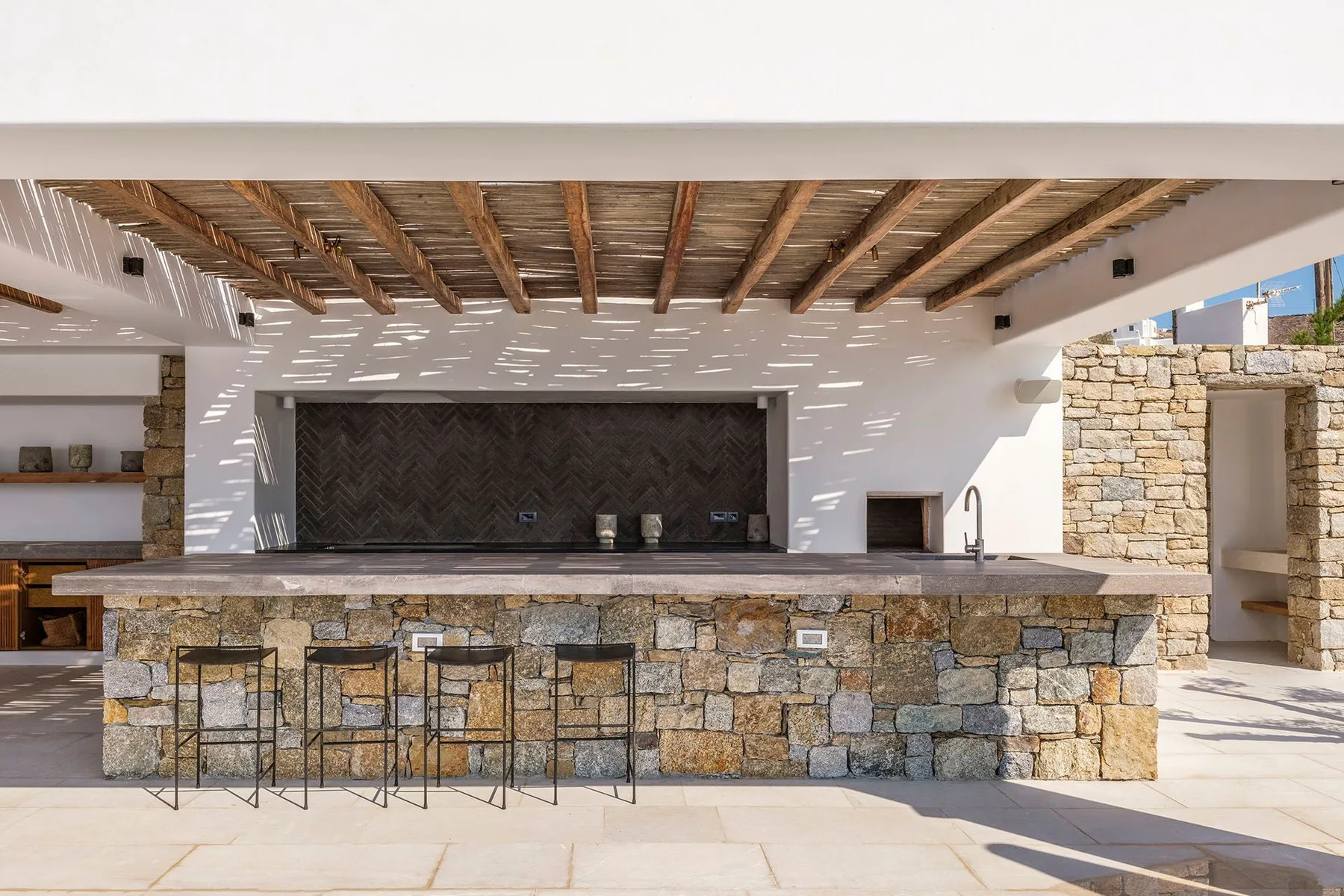An outdoor bar with a stone counter and stools beneath a wooden-beamed roof.