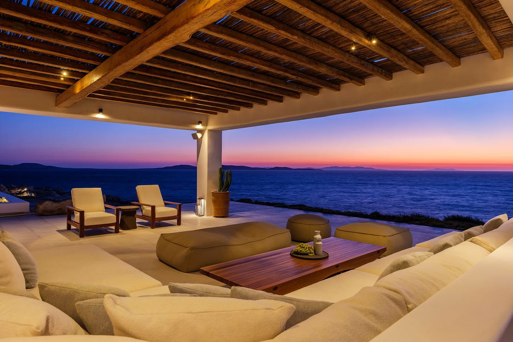 An open-air lounge overlooks the ocean at sunset, with cushioned seating beneath a wooden ceiling.