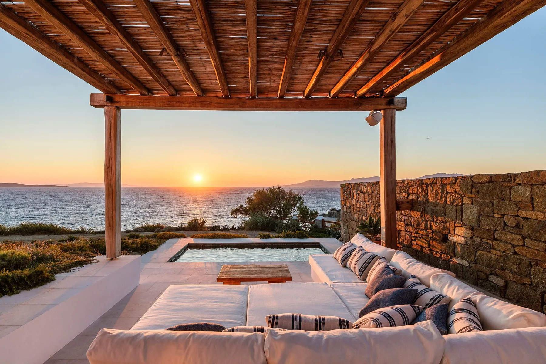 A seaside patio with cushioned seating overlooks the water at sunset.