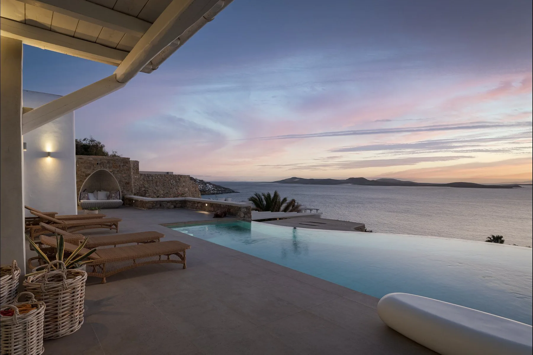 Una piscina infinita junto a una zona de estar exterior da al mar al atardecer.