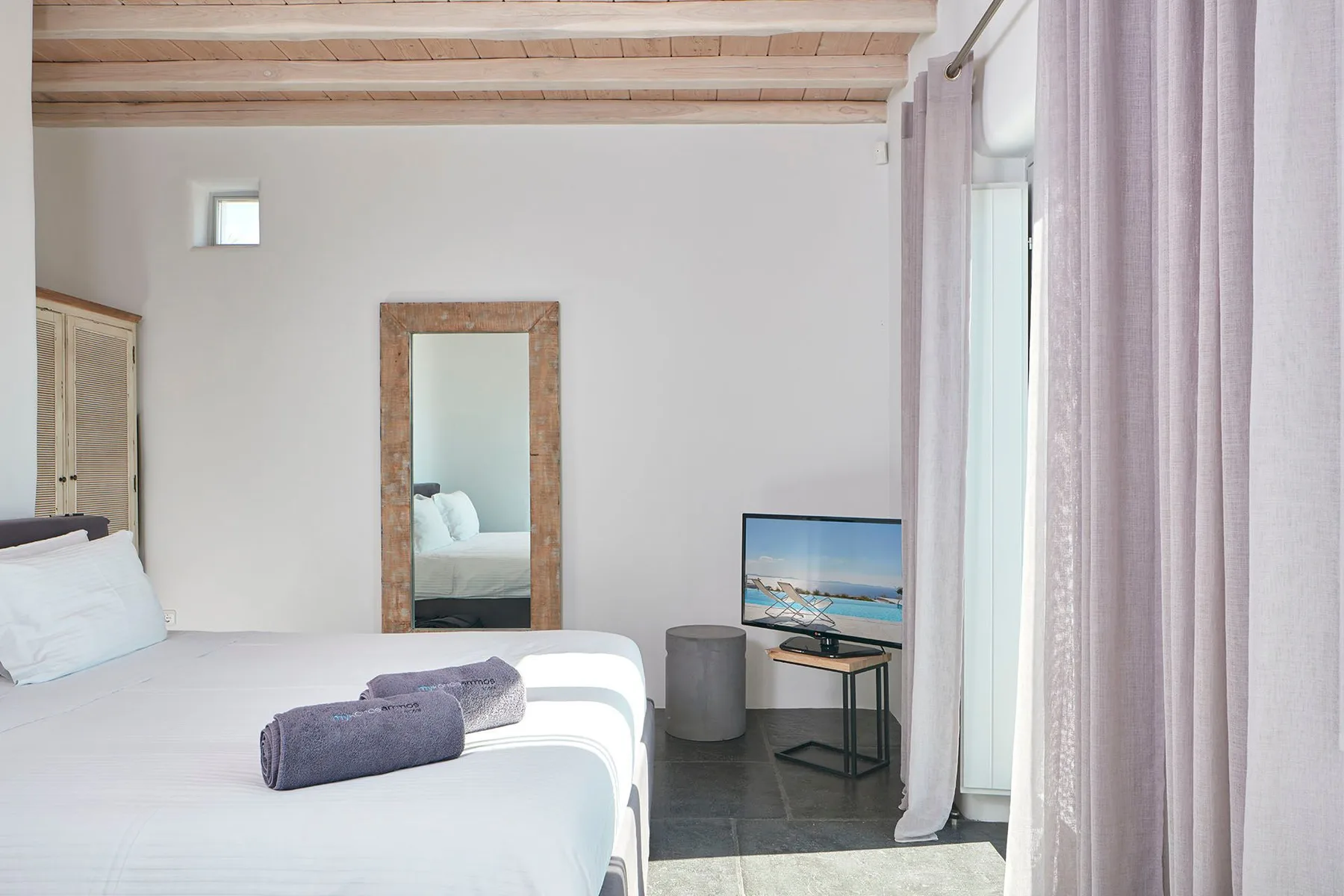 A bedroom with white bedding, a mirror, and curtains beside a window.