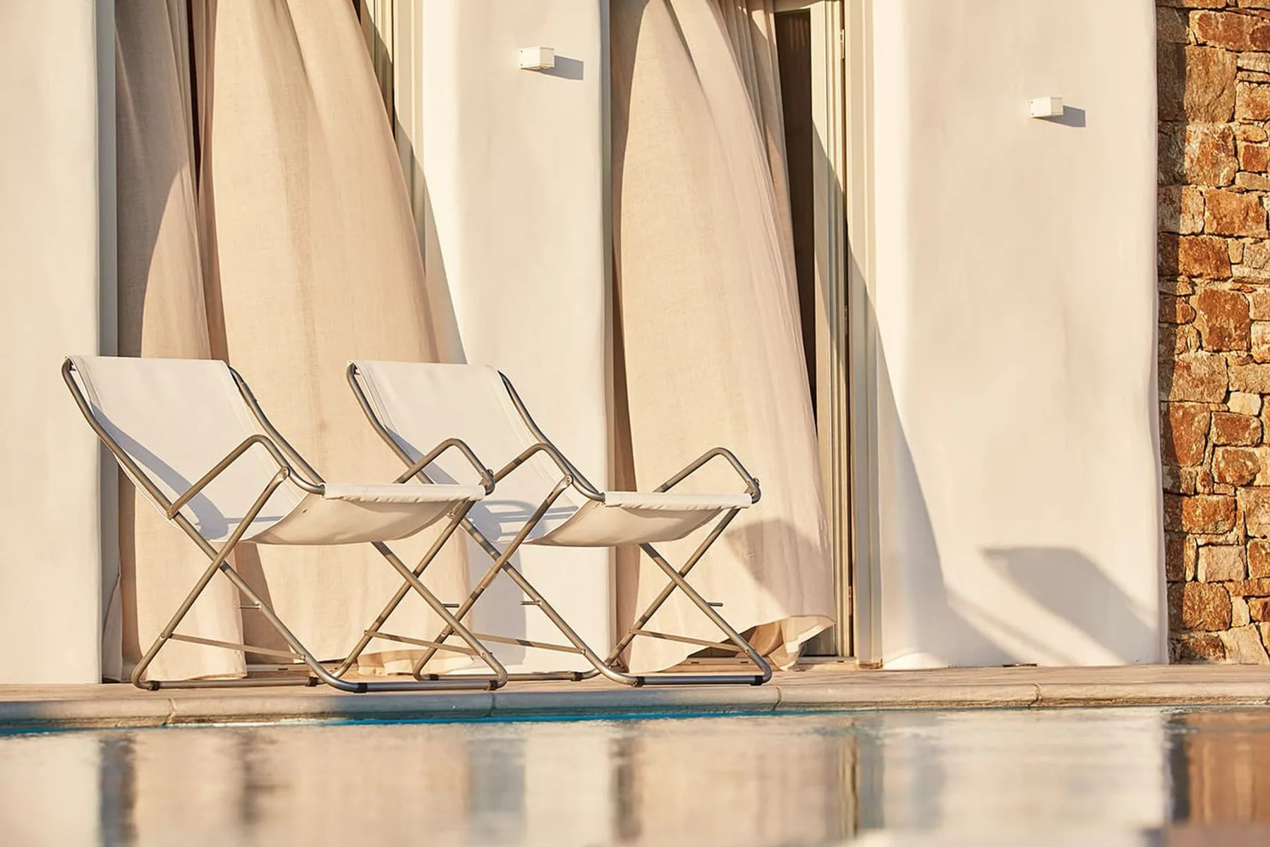 Outdoor lounge seating sits beside a pool, with pale curtains along the wall.