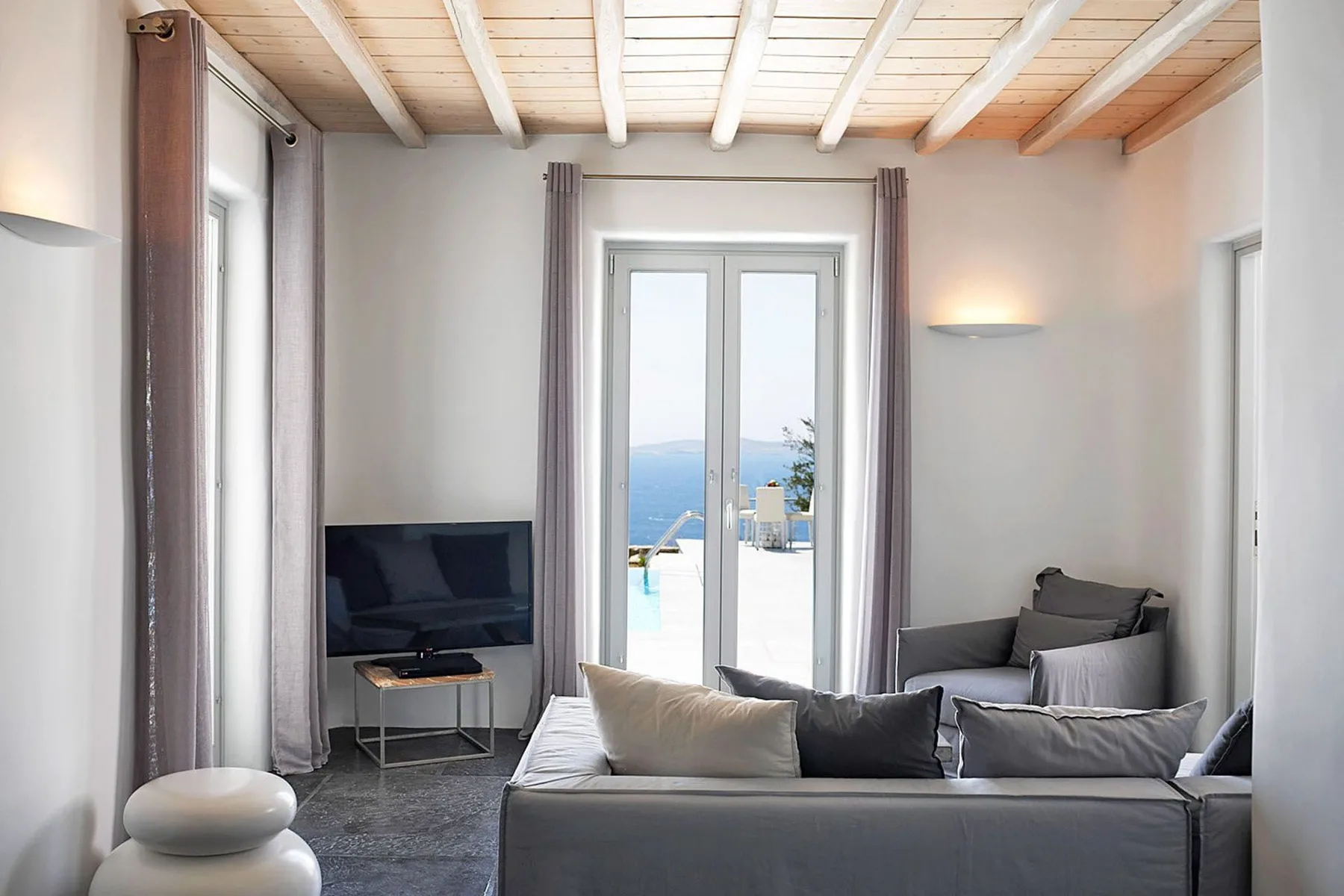 A bright living room with seating, a television, and doors opening toward a pool and sea view.